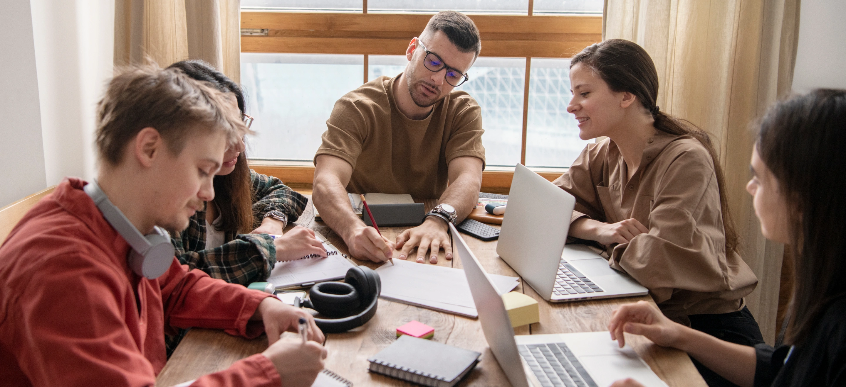 Five young adults working together at a wooden table with laptops, notebooks, and headphones near a window.