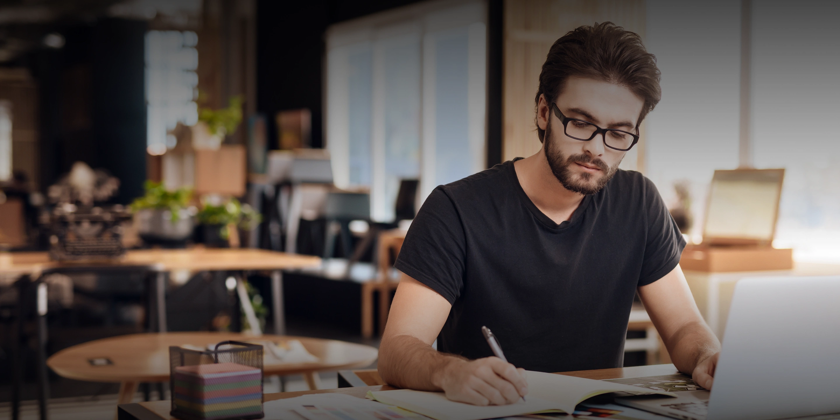 Man wearing glasses and a black t-shirt writing in a notebook at a wooden table with a laptop, in a modern office setting.
