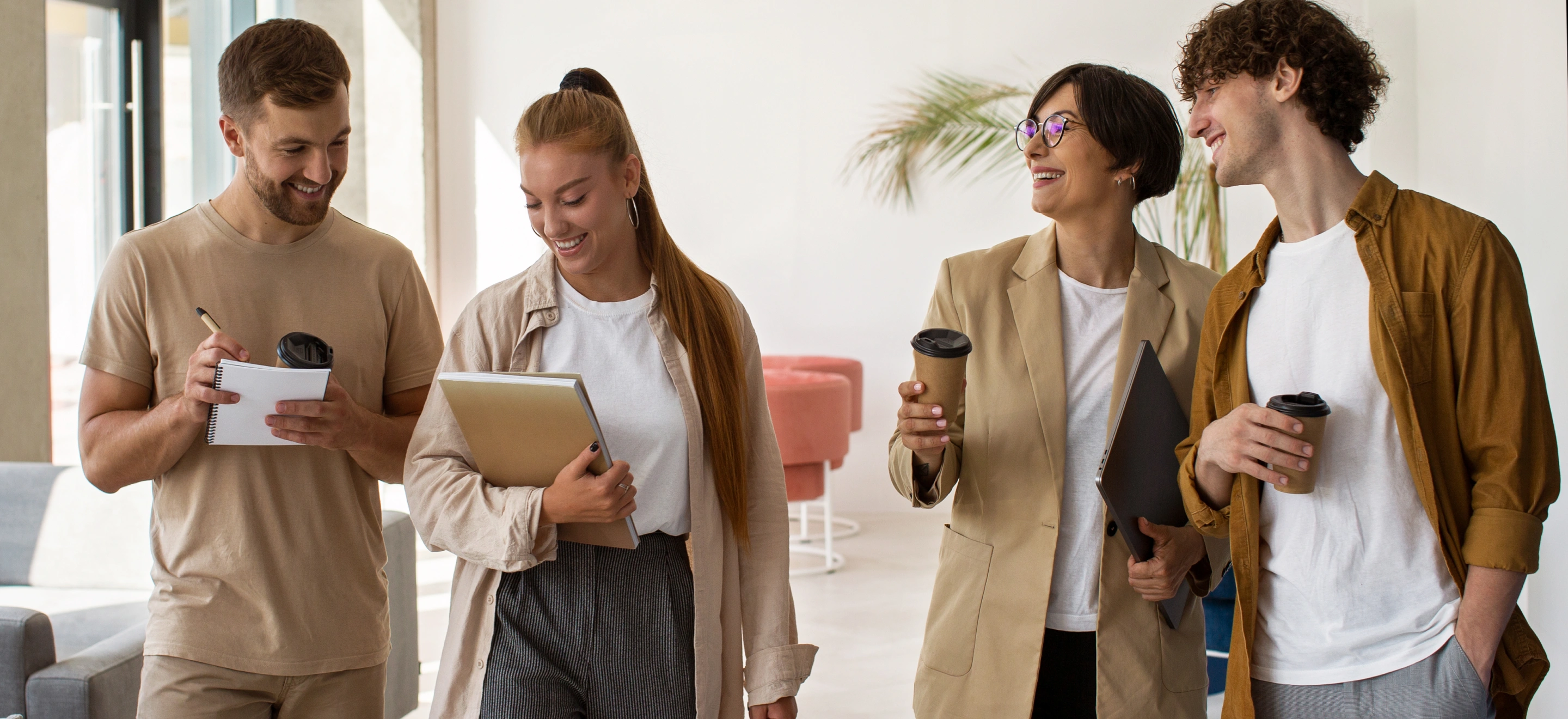 Four young adults casually dressed, smiling and interacting while holding notebooks, a laptop, and coffee cups in a bright indoor space.