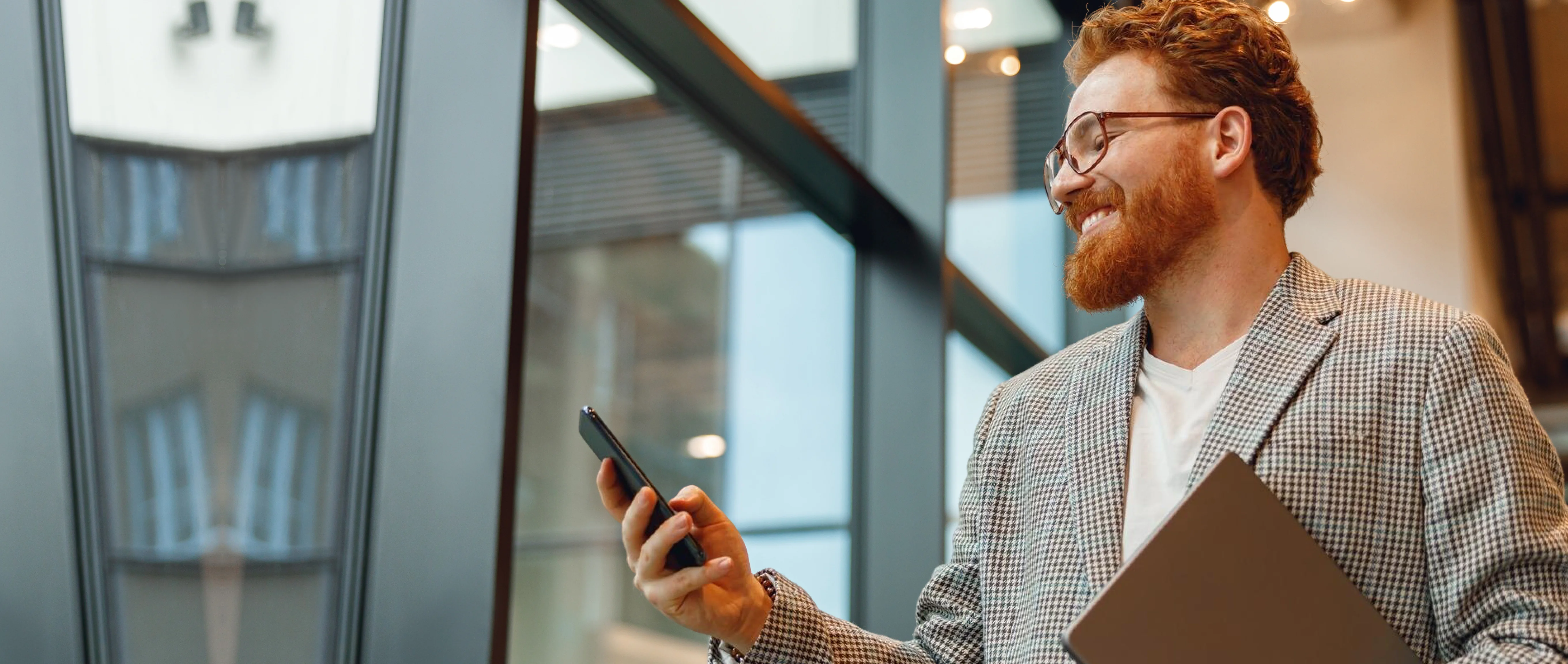 Smiling man with glasses and a beard holding a laptop and looking at his smartphone near large windows in an office.