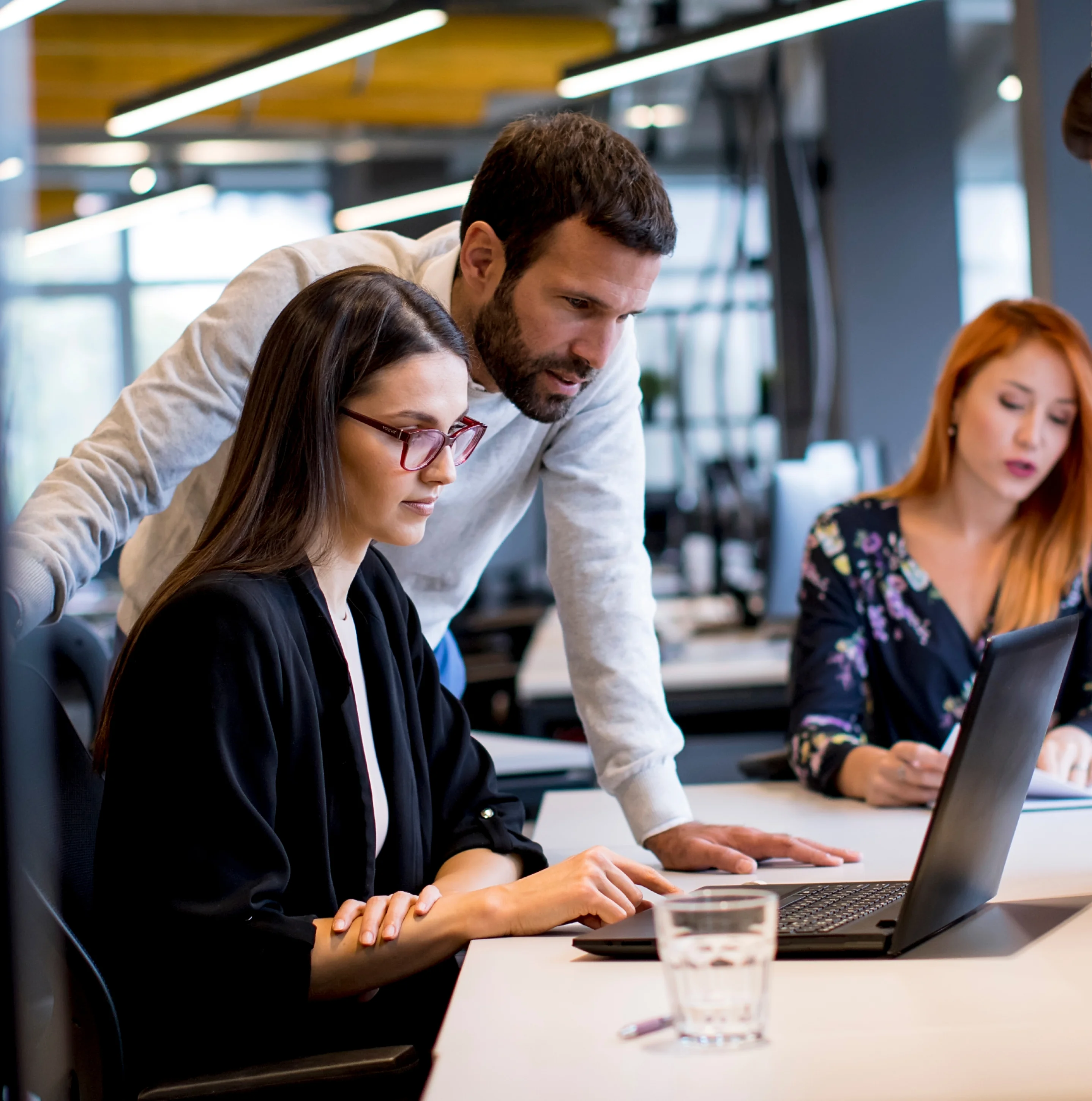 Due colleghi concentrati che lavorano su un laptop in un ufficio moderno, con una terza persona sullo sfondo.
