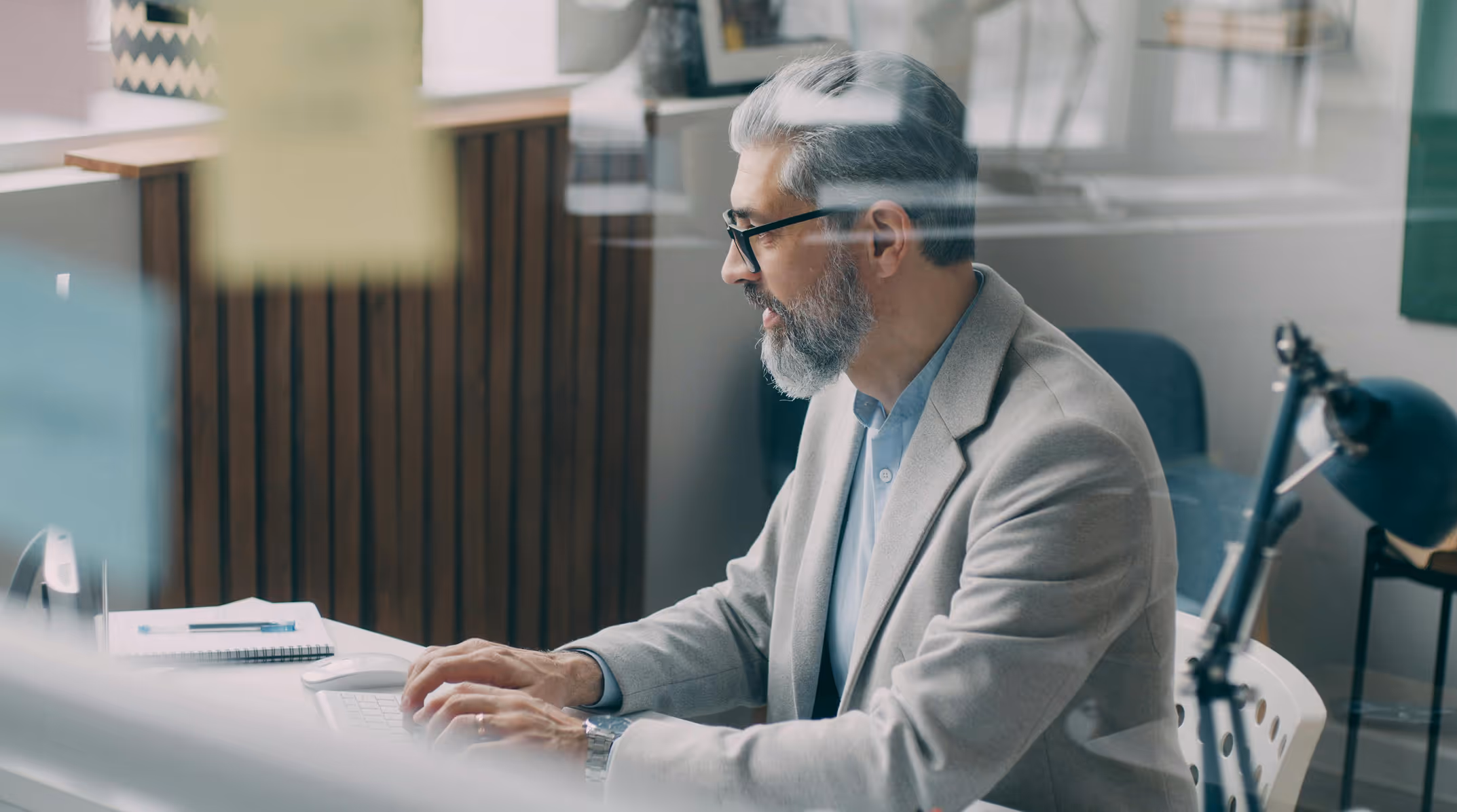 Senior man with gray hair and beard, wearing glasses and a light gray blazer, working on a computer in an office setting.