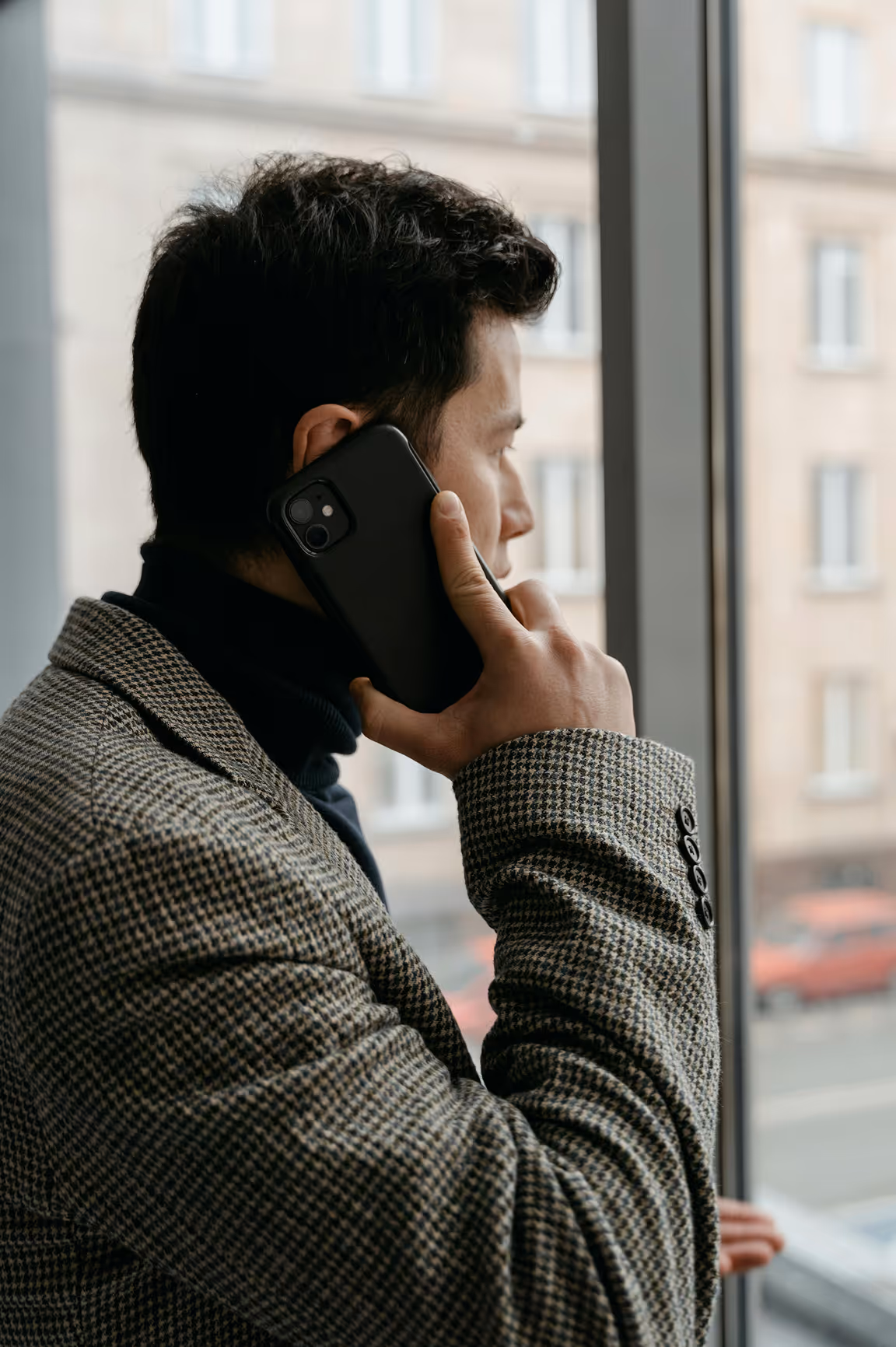 Man in a checkered jacket talking on a smartphone while looking out of a window.