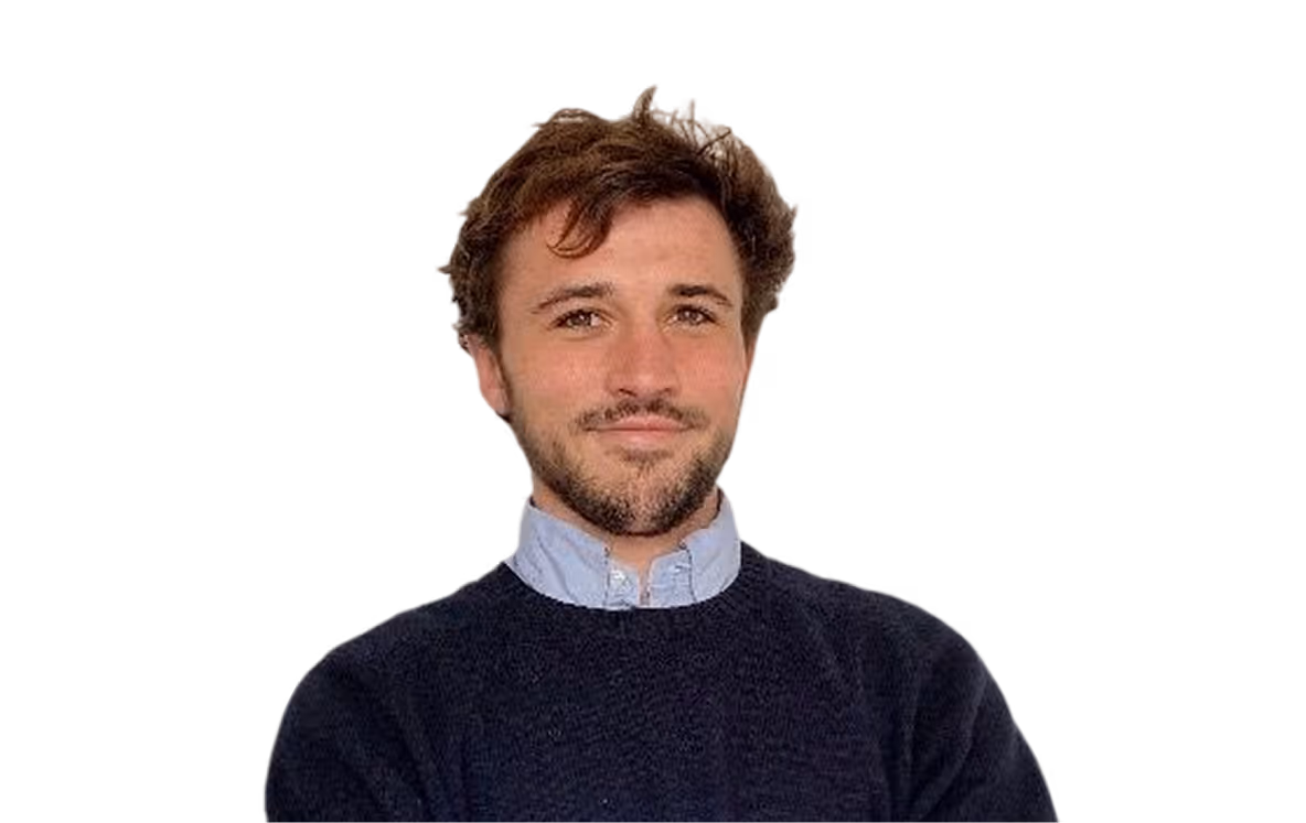 Smiling young man with short curly brown hair and a beard wearing a blue sweater over a light blue collared shirt against a pale background, the man is Ambroise COUCHET.