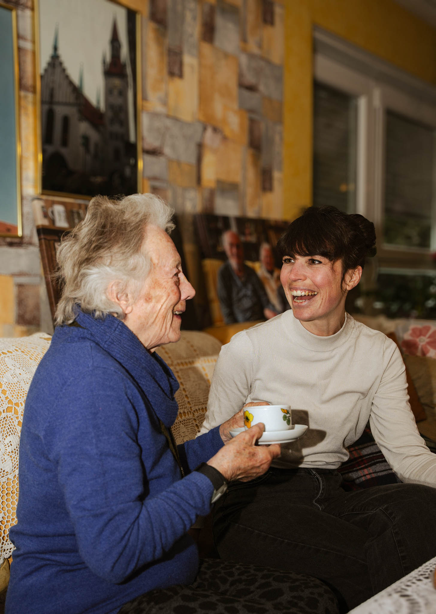 Ältere Frau in blauem Pullover trinkt Kaffee und unterhält sich lachend mit jüngerer Frau.