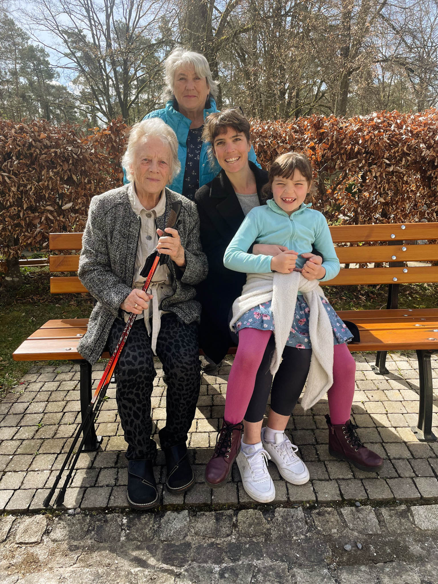 Vier Generationen Frauen sitzen auf einer Parkbank im Freien, darunter eine ältere Frau mit Gehhilfen, eine jüngere Frau und ein Mädchen.