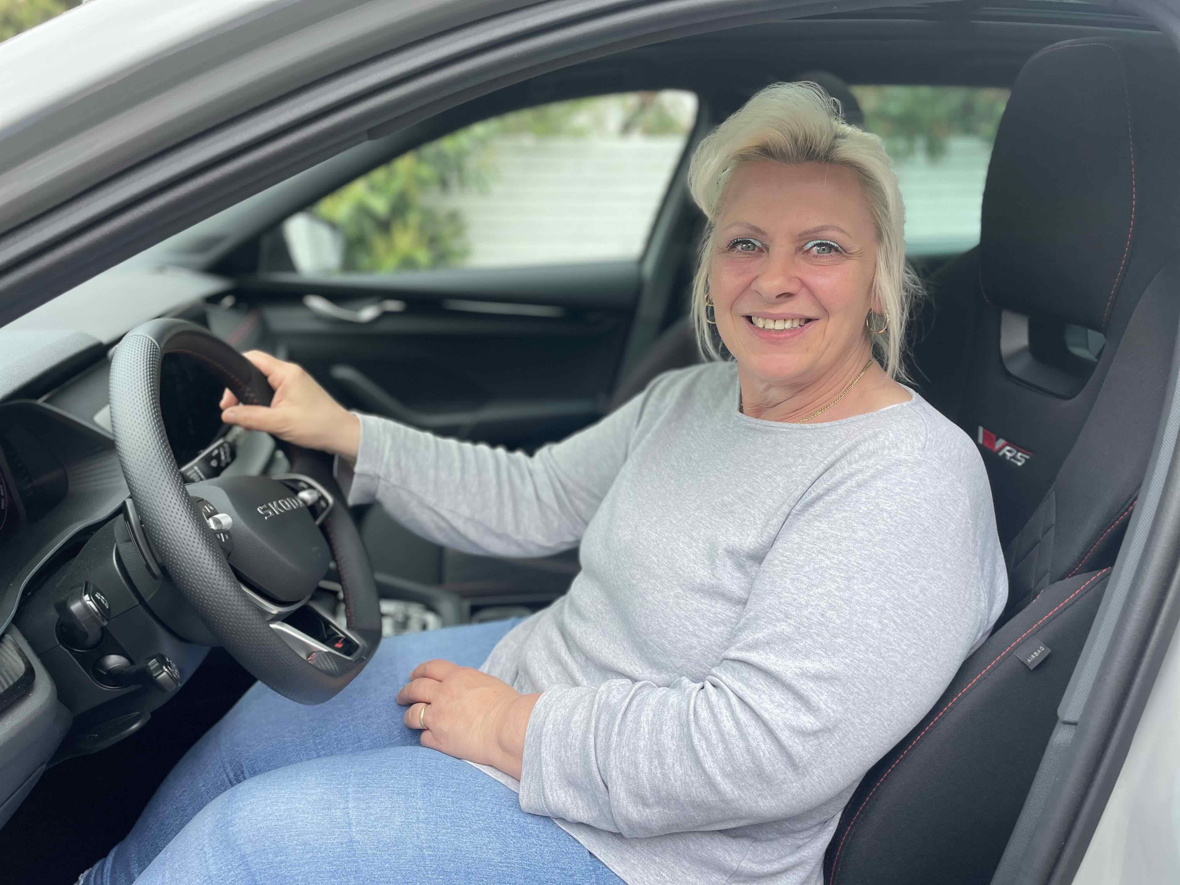 Frau mit blonden Haaren sitzt lächelnd am Steuer eines Fahrzeugs.