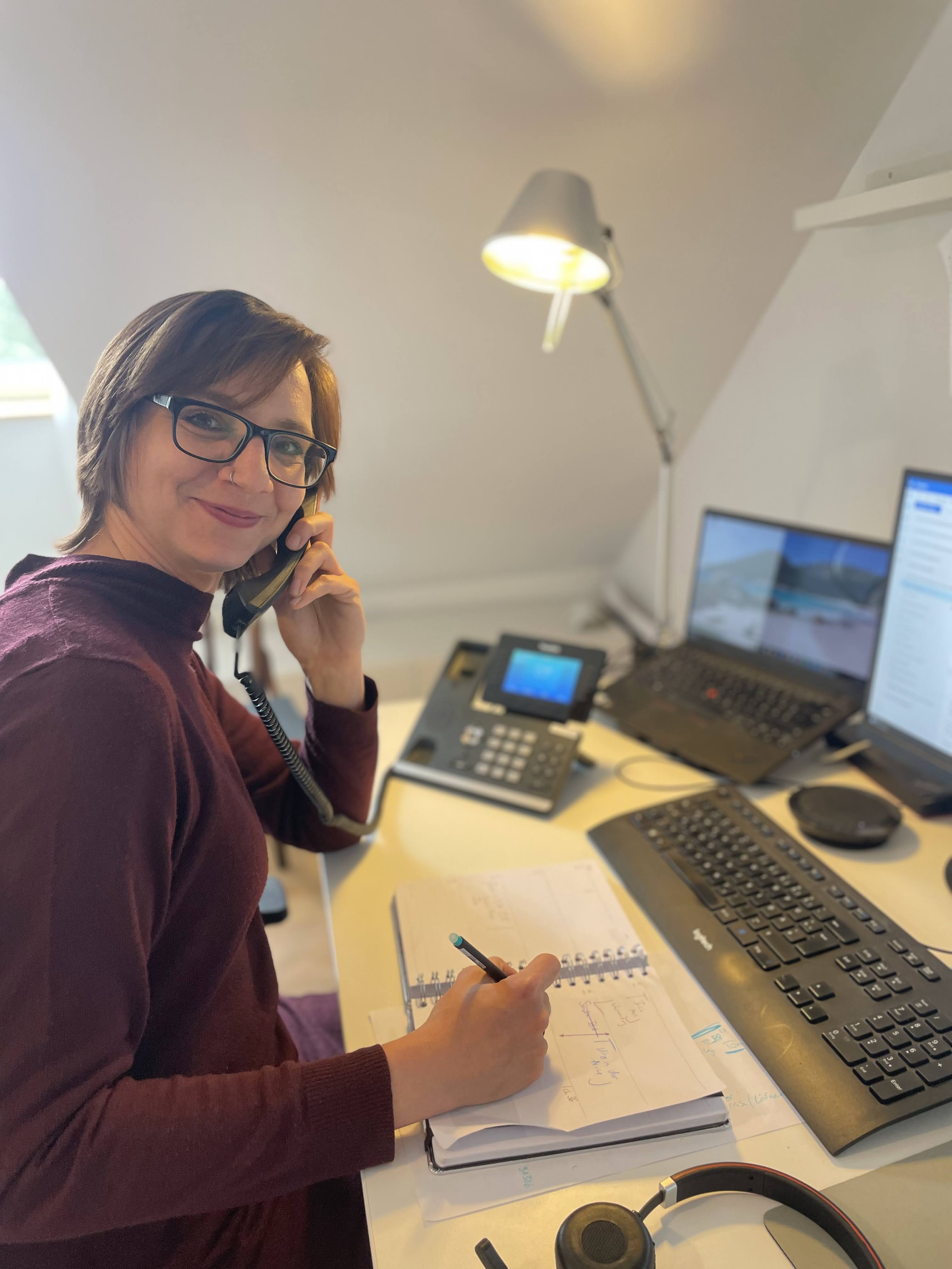 Frau telefoniert am Schreibtisch und macht Notizen am Laptop mit drei Bildschirmen.