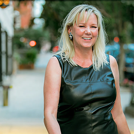 Smiling young woman with long blonde hair, blue eyes, and hoop earrings wearing a beige turtleneck and navy blazer.