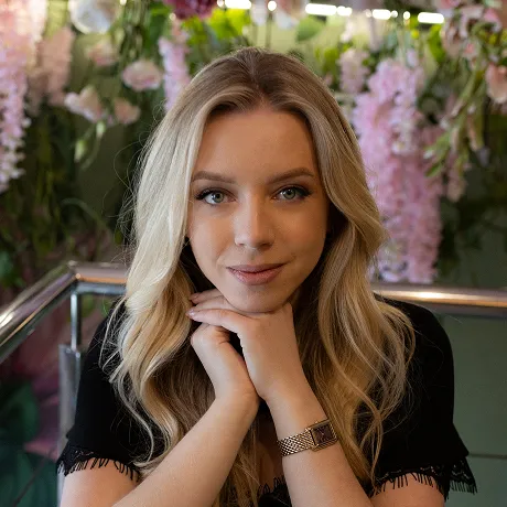 Smiling young woman with long blonde hair, blue eyes, and hoop earrings wearing a beige turtleneck and navy blazer.