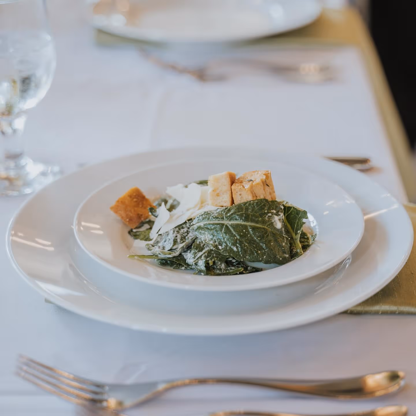 White plate with Caesar salad featuring romaine lettuce, croutons, and shaved Parmesan cheese on a white tablecloth with silverware.