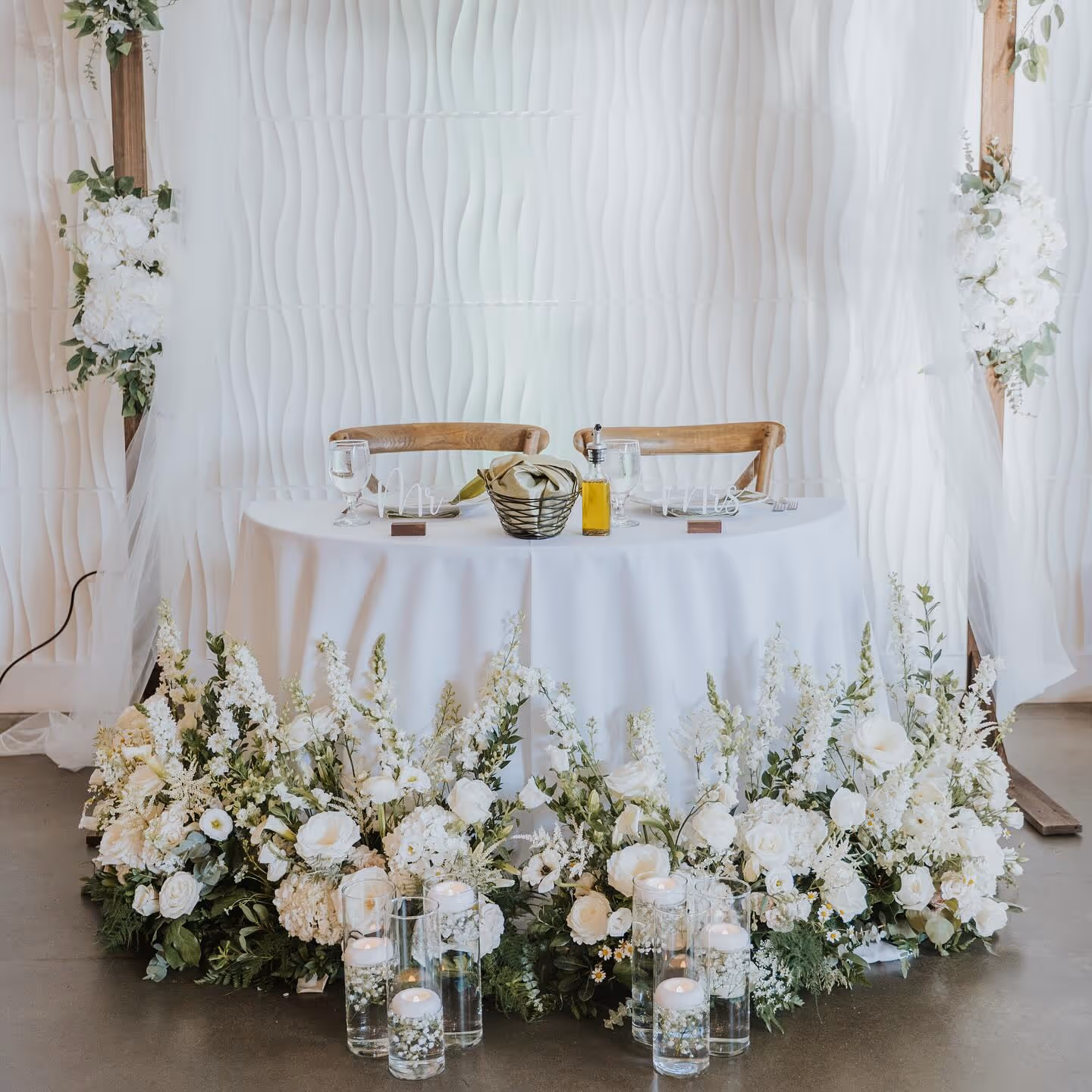 Wedding sweetheart table set with white tablecloth, two wooden chairs, white floral arrangements, and glass candles in cylindrical holders.