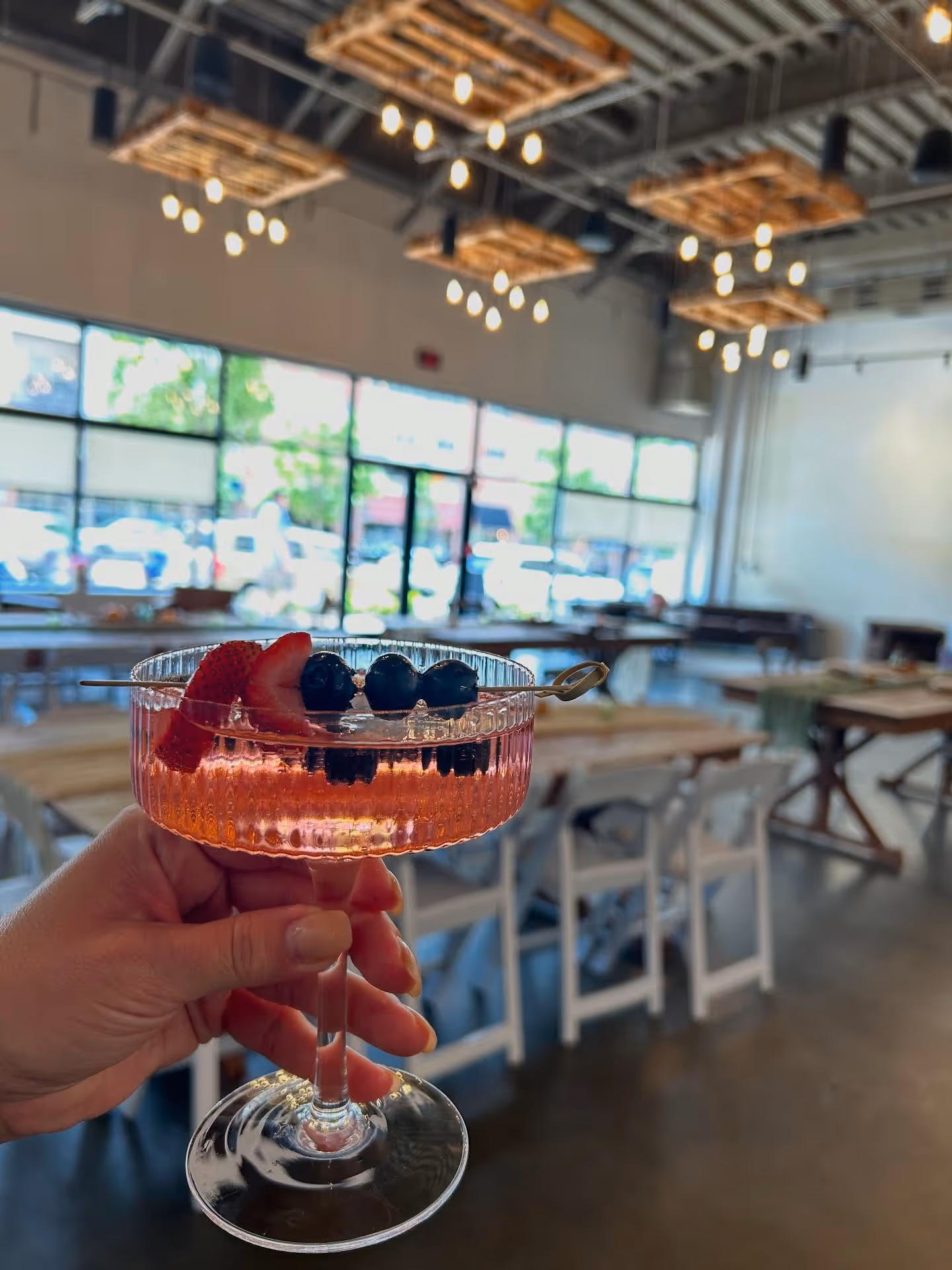 Hand holding a pink cocktail garnished with a strawberry slice and blueberries on a skewer in a bright, modern dining space with wooden tables and white chairs.