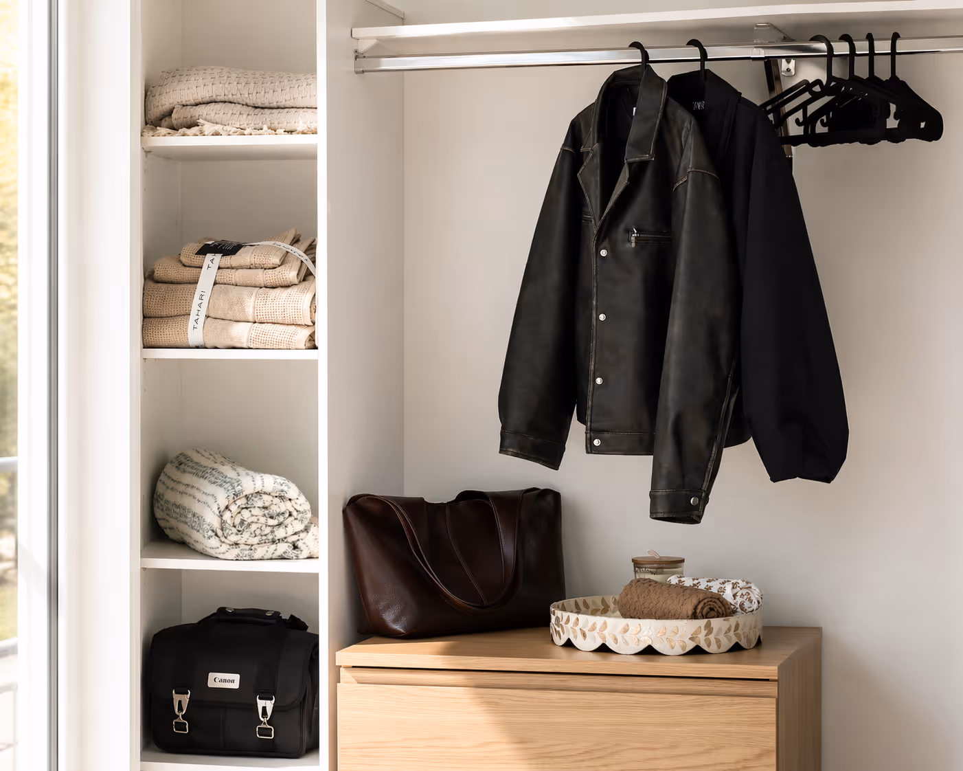 Closet with black leather and fabric jackets hanging, folded towels on shelves, and bags on dresser top.