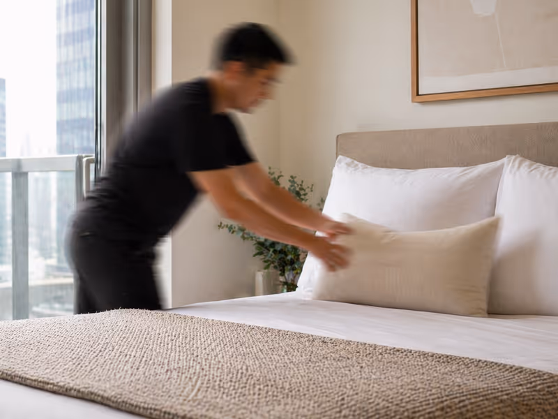 Person arranging a beige pillow on a neatly made bed with white pillows and a textured blanket.