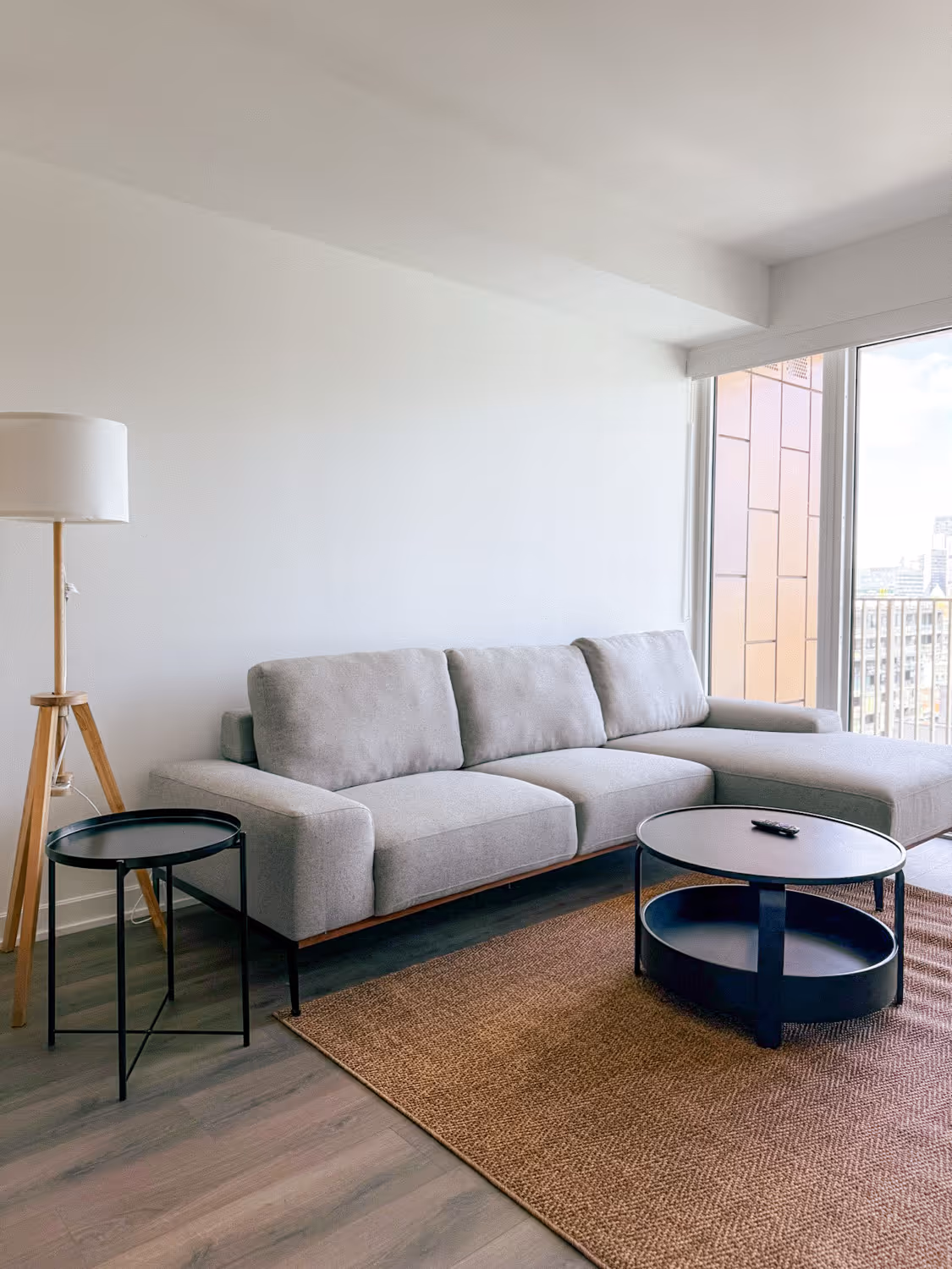 Modern living room with gray sectional sofa, round black coffee table, tall wood lamp, and large window.