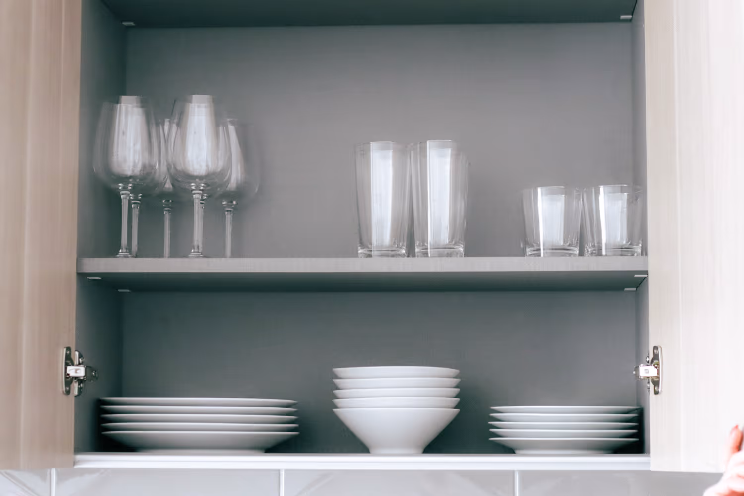 Kitchen cabinet with stacked white plates and bowls on the bottom shelf and clear glasses on the top shelf.