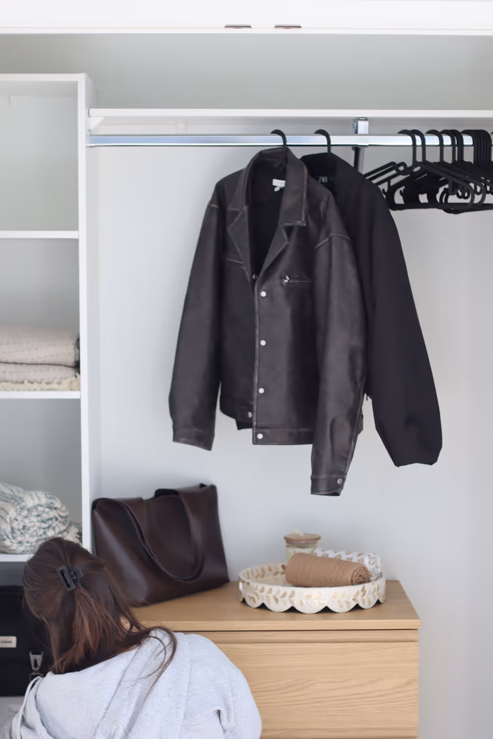 Black jackets hanging in a closet with folded clothes, a leather bag, and a woman organizing clothes below.