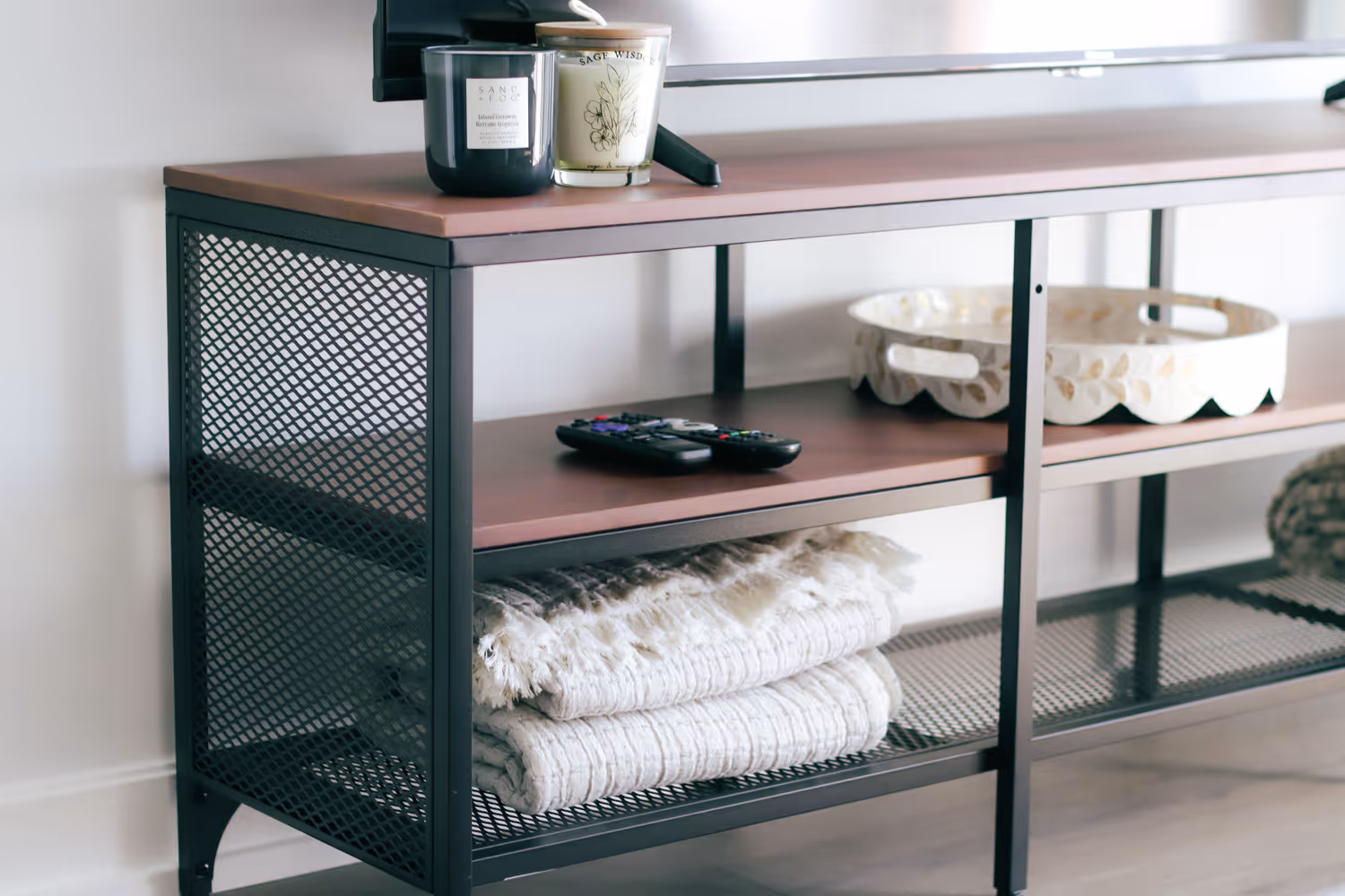 Metal and wood TV stand with two candles, remote controls, white tray, and folded textured blankets.