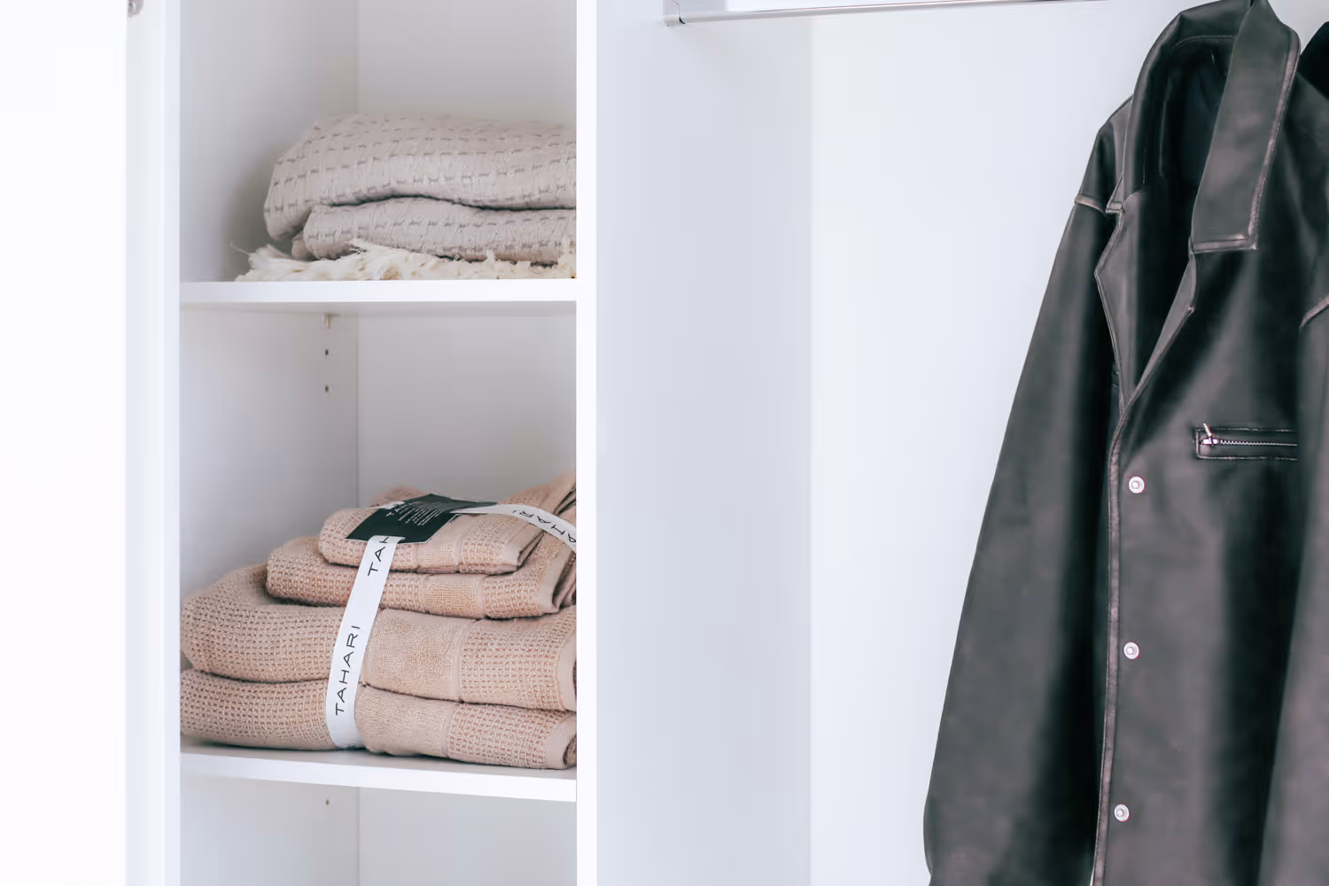 Beige folded towels on shelves next to a hanging black leather jacket in a white closet.