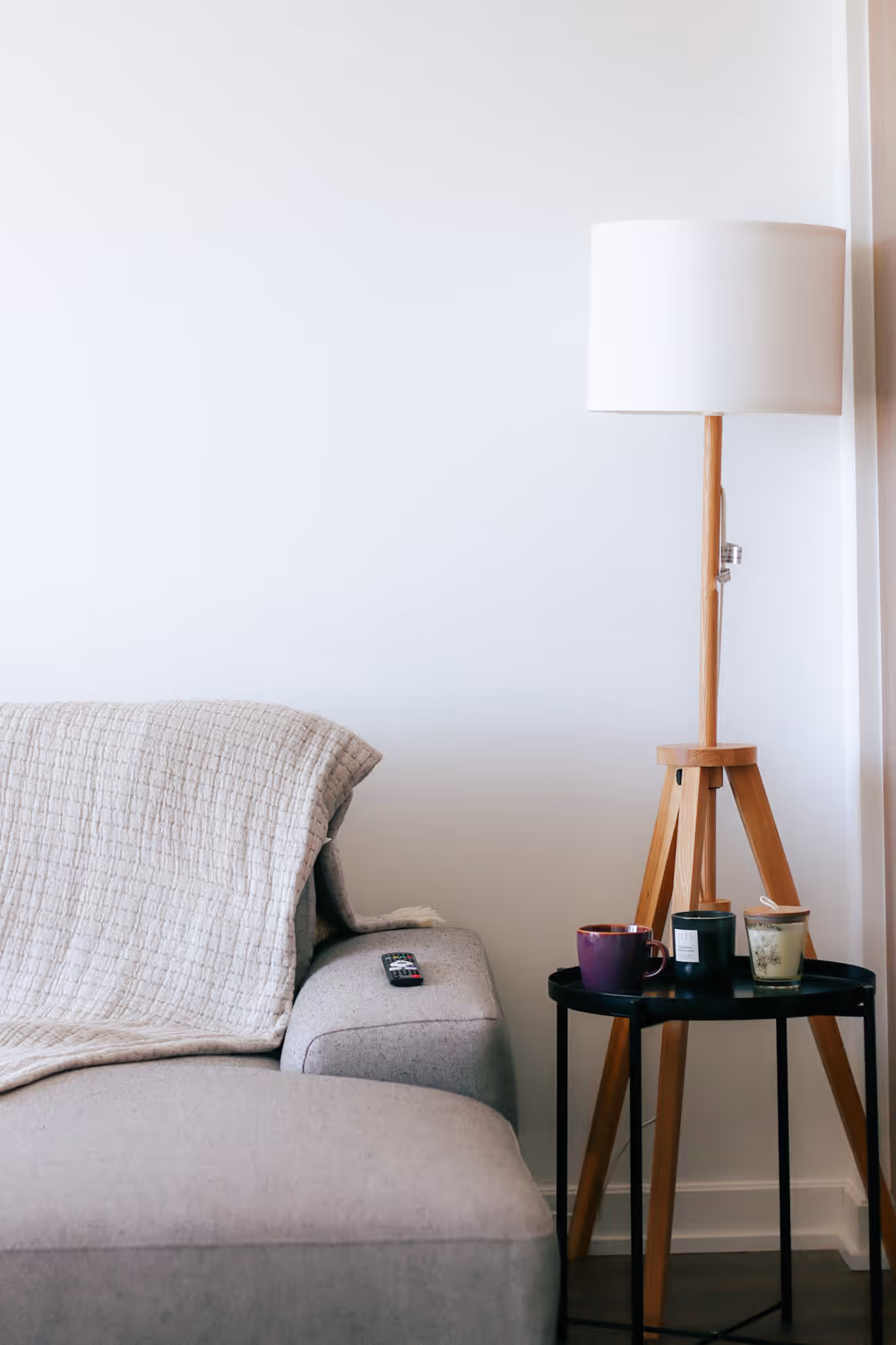 Gray sofa with a remote, beige throw, next to a wooden tripod lamp and a black table with cups and candles.