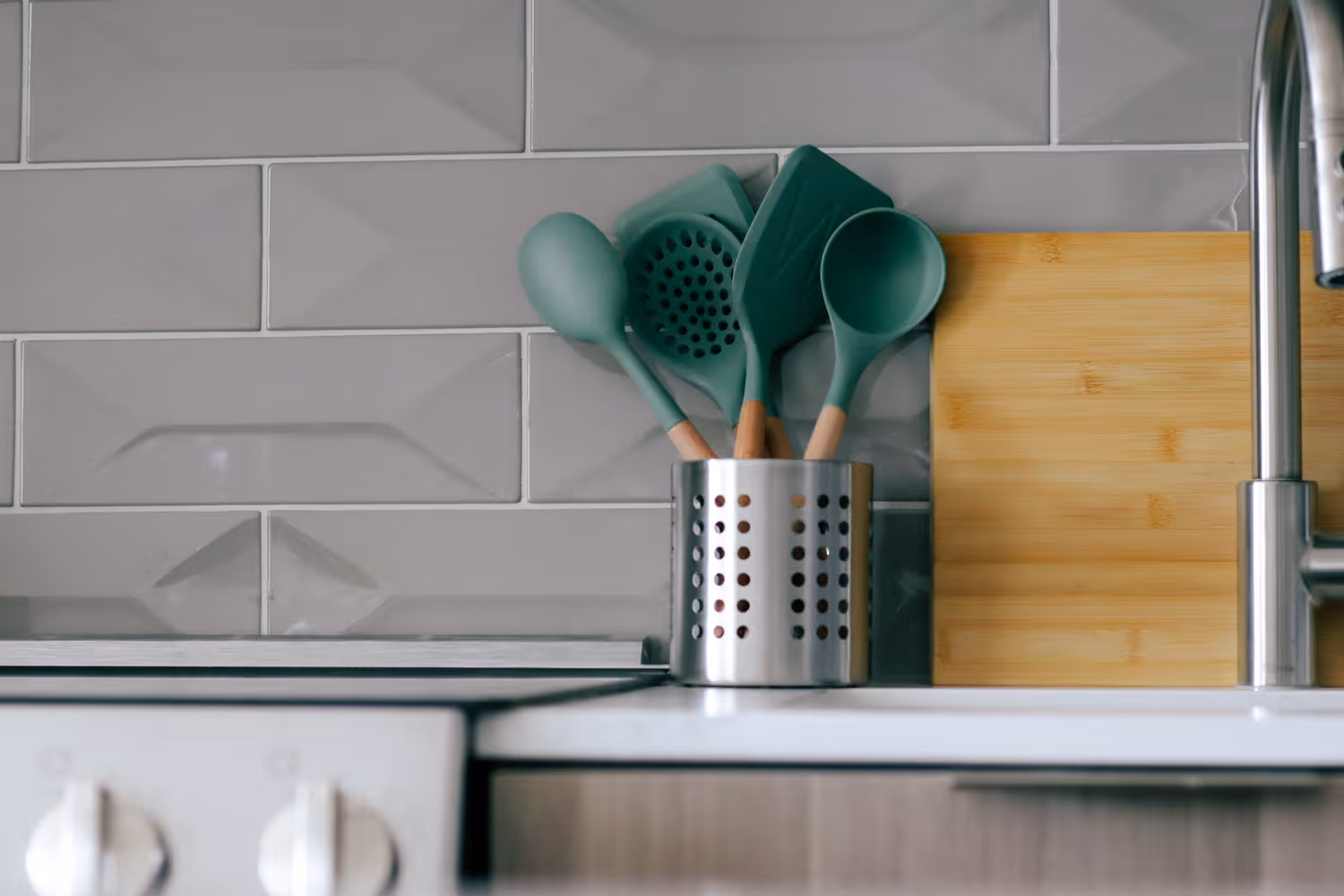 Green kitchen utensils in a metal holder beside a bamboo cutting board and stainless steel faucet.