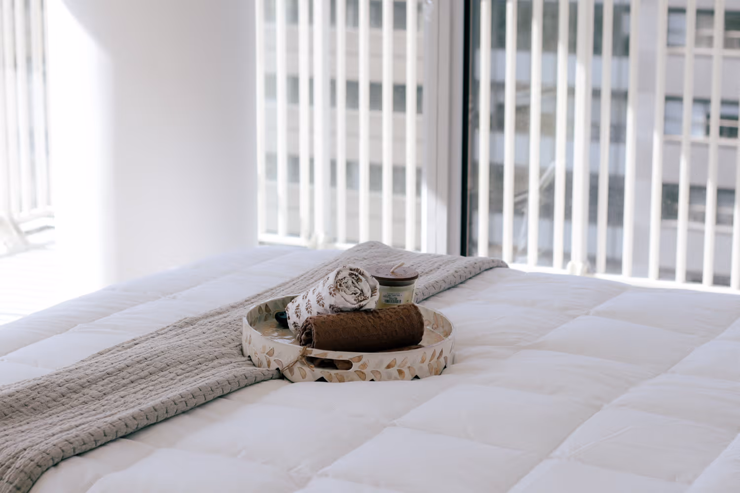 Tray with rolled towels and a candle on a neatly made white bed with a beige runner near a window.