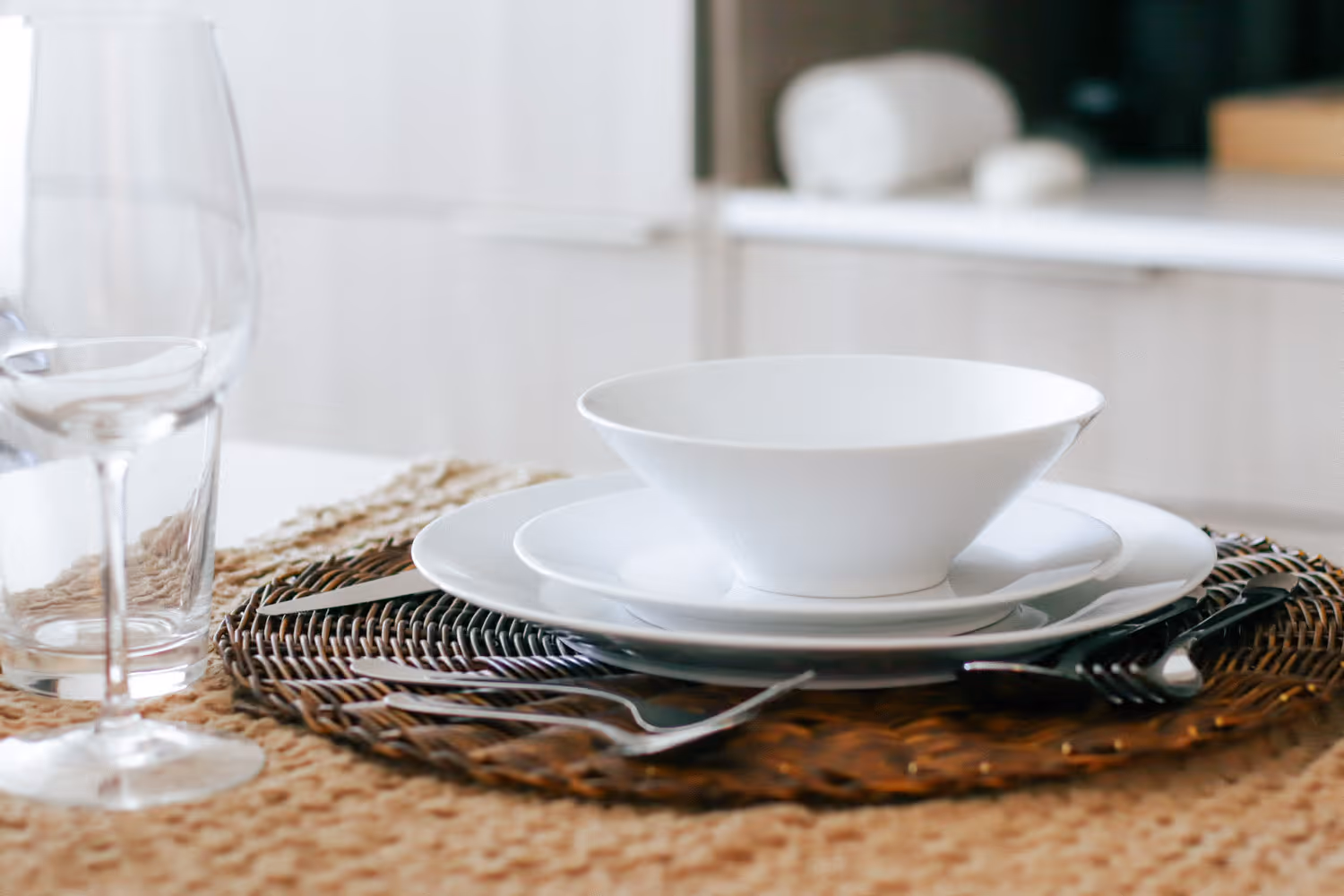 White bowl on stacked white plates with silverware and a clear wine glass on woven placemat.