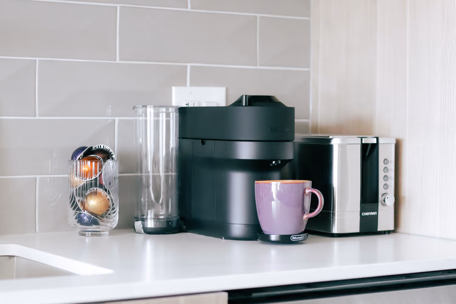 Black coffee machine with purple mug and clear container of capsules on white kitchen counter.