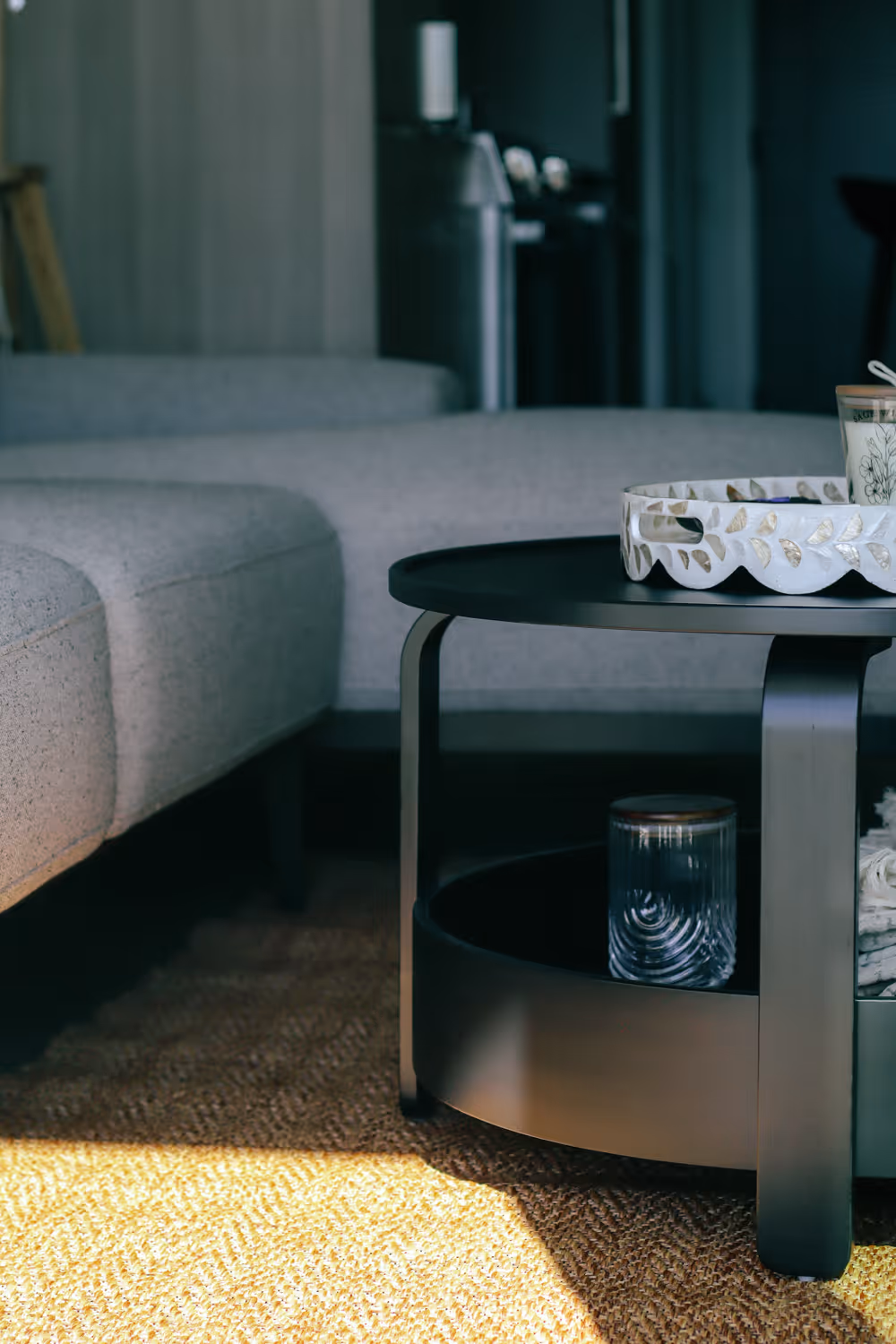 Gray sofa beside a black round coffee table with a decorative tray and glass jar on a woven rug.