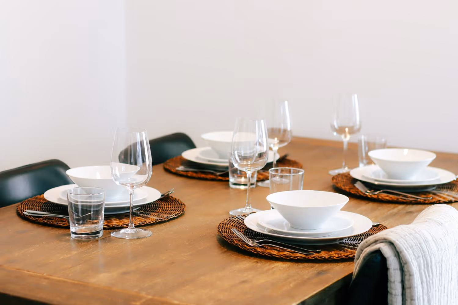 Wooden dining table set with white bowls, plates, wine glasses, and woven placemats.
