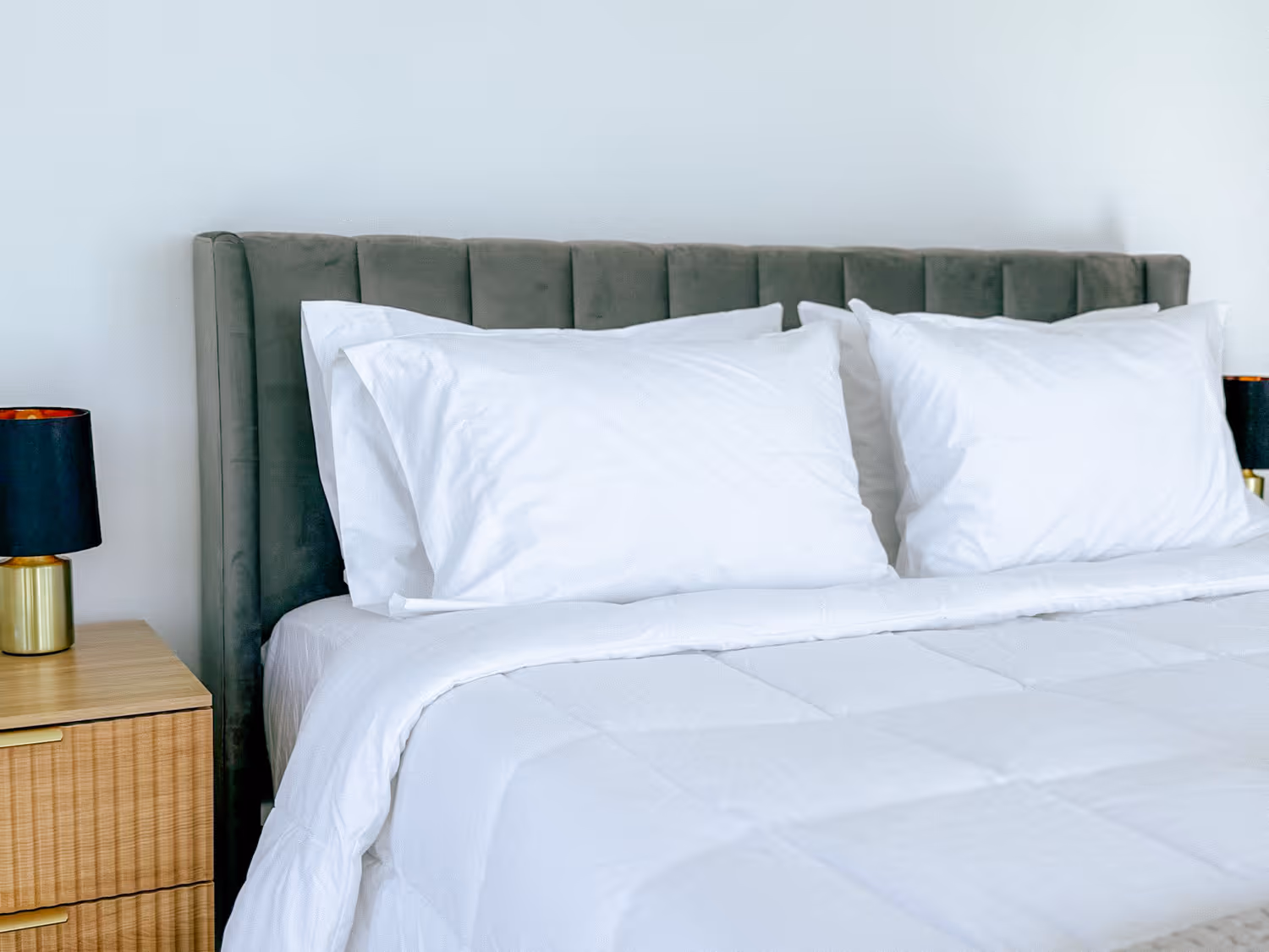 Bed with white pillows and comforter, gray upholstered headboard, and wooden nightstand with black lamp.