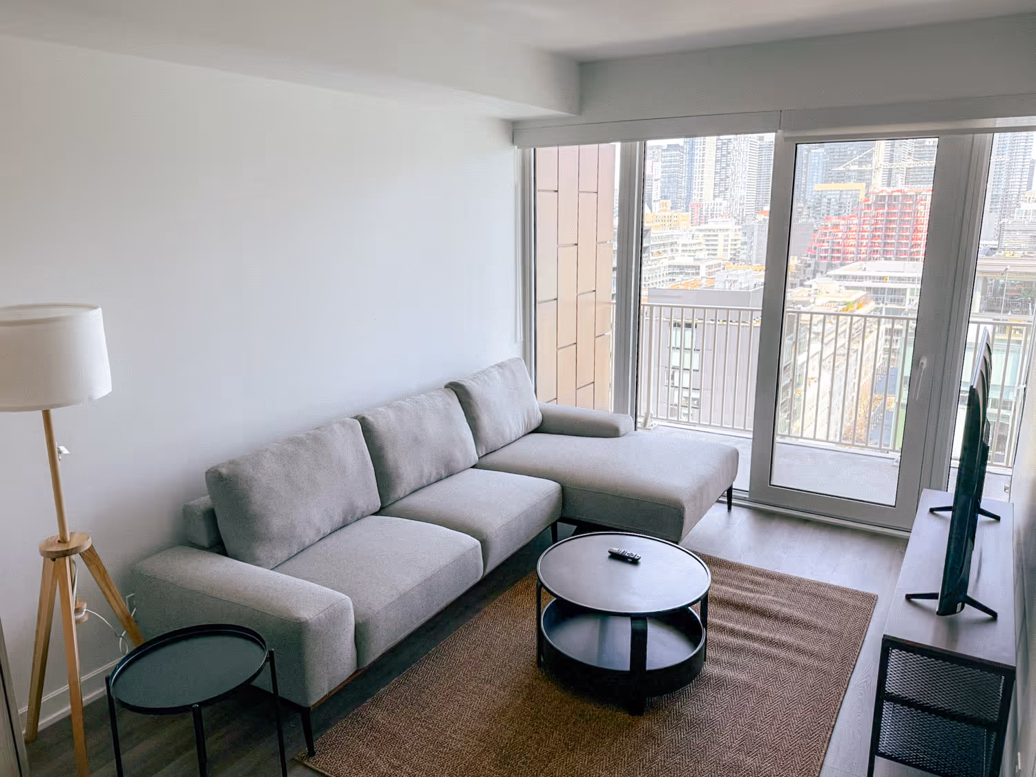 Modern living room with gray sectional sofa, round black coffee table, floor lamp, and city view balcony.