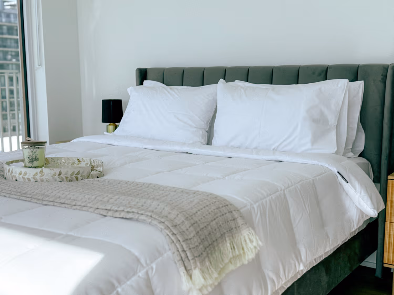 Modern bed with white bedding, pillows, beige throw, and a tray with candle by a balcony window.