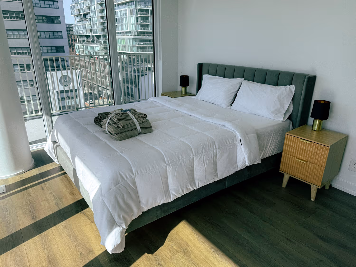 Modern bedroom with white bed linens, green headboard, folded towels, wood floor, and city view window.