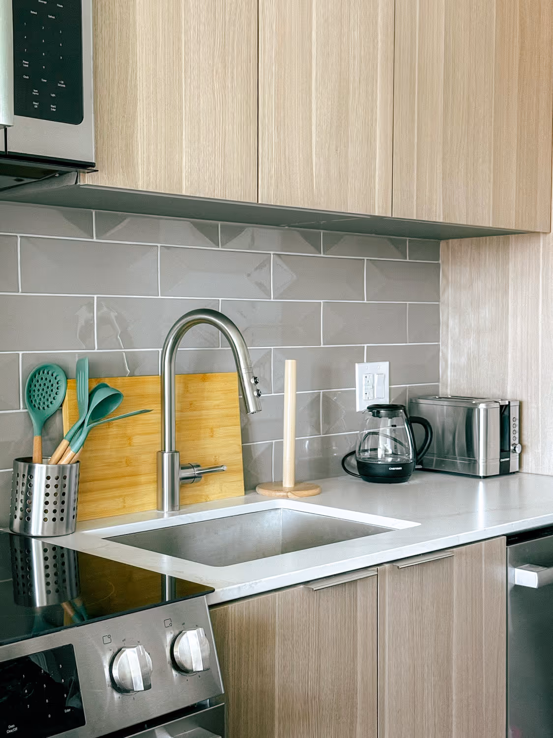Modern kitchen sink with stainless faucet, utensils, coffee maker, toaster, and stovetop controls.