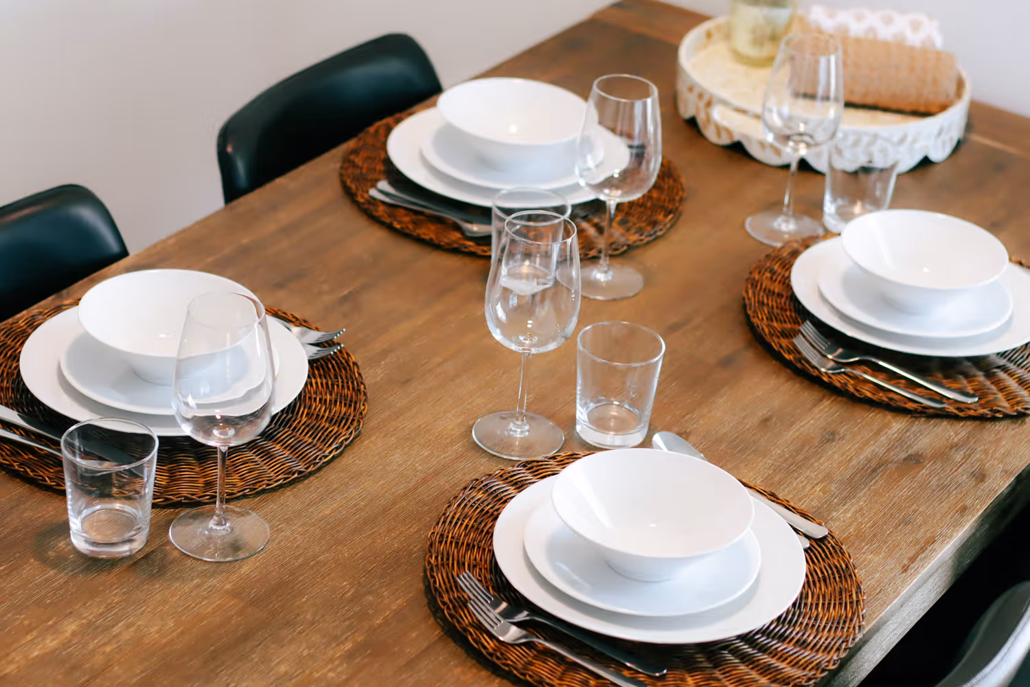 Wooden dining table set for four with white dishes, woven placemats, wine glasses, and silverware.