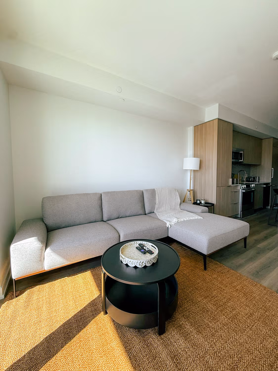 Modern living room with gray sectional sofa, black coffee table, lamp, and kitchen in background.
