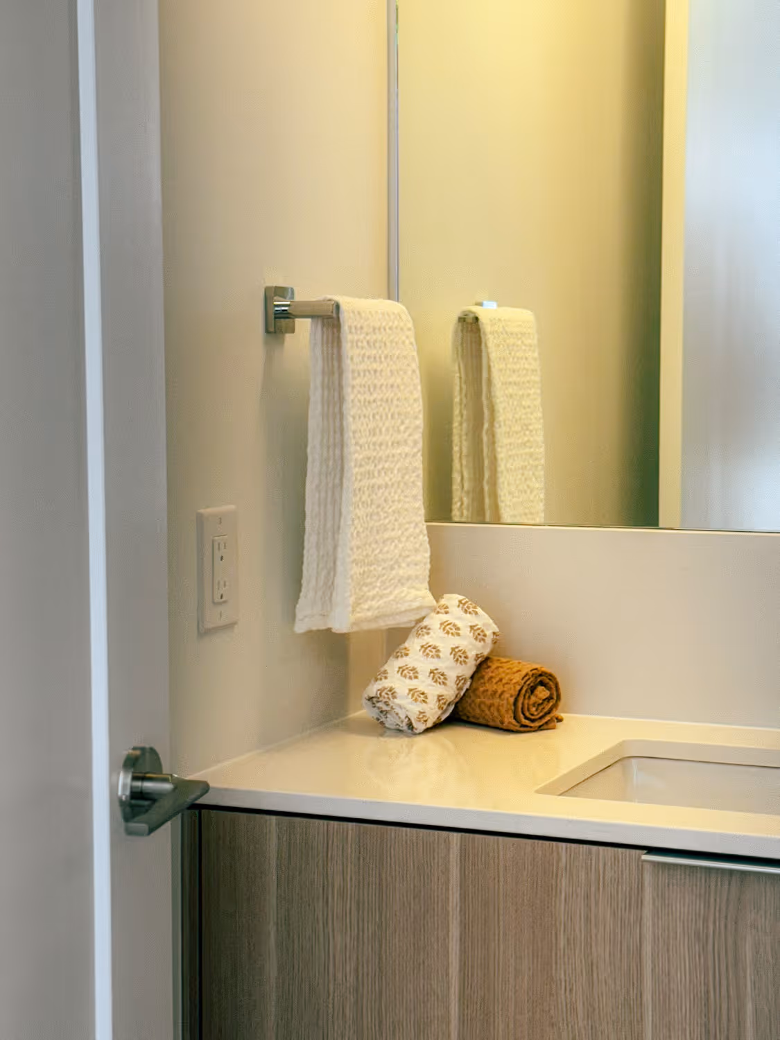 White bathroom countertop with rolled towels and a hanging knitted white towel beside a mirror.