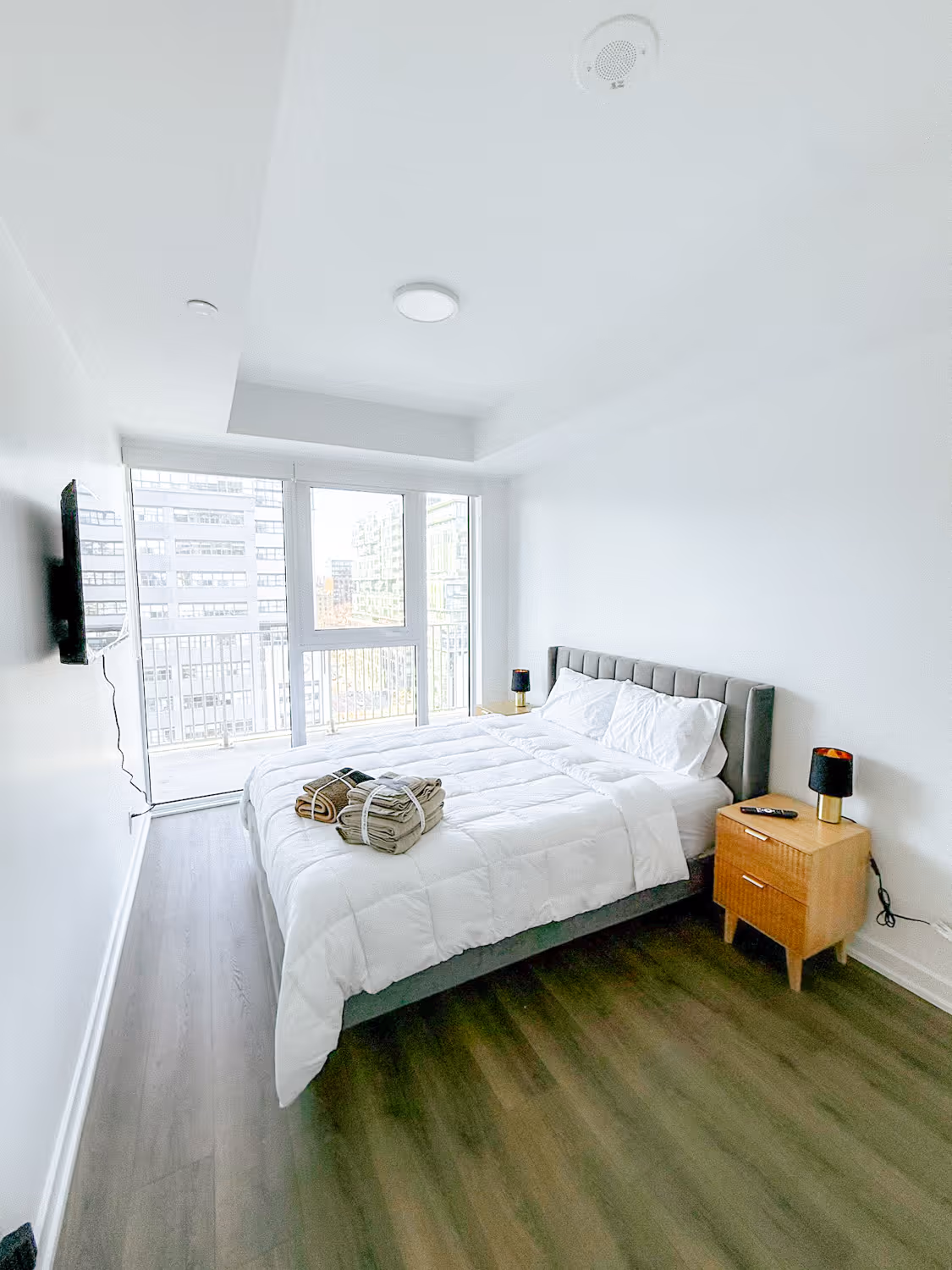 Bright bedroom with large window, white bedding on queen bed, wooden nightstand, and mounted TV.