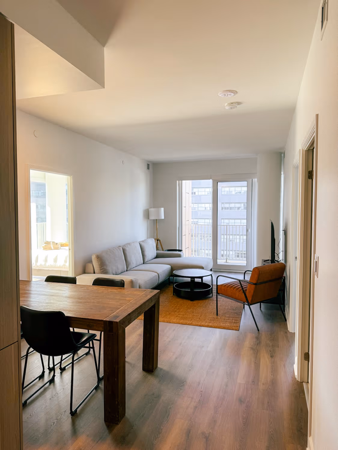 Modern living room with gray sofa, orange armchair, wooden dining table, and large window balcony doors.