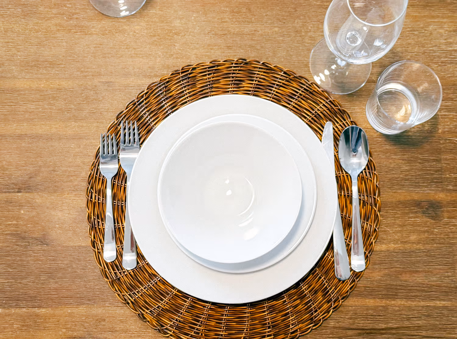 Table setting with white plates on a wicker charger, two forks, knife, spoon, wine glass, and water glass.