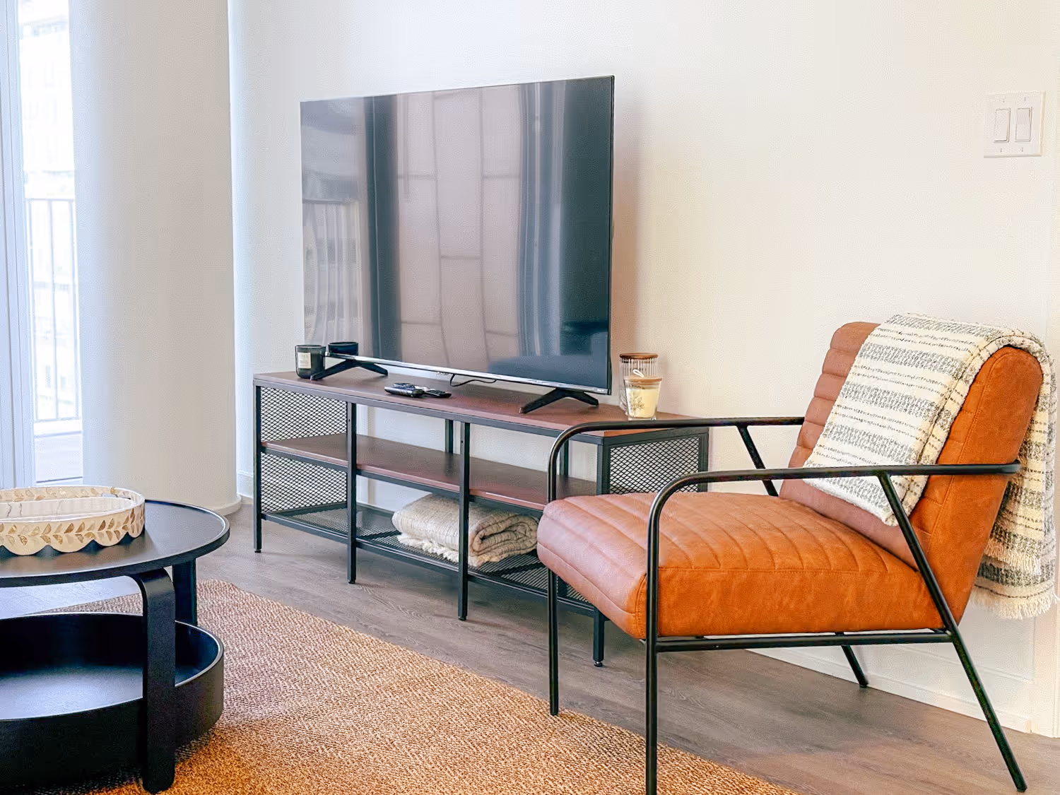 Modern living room with a large flat-screen TV on a metal and wood stand and an orange armchair with a striped blanket.