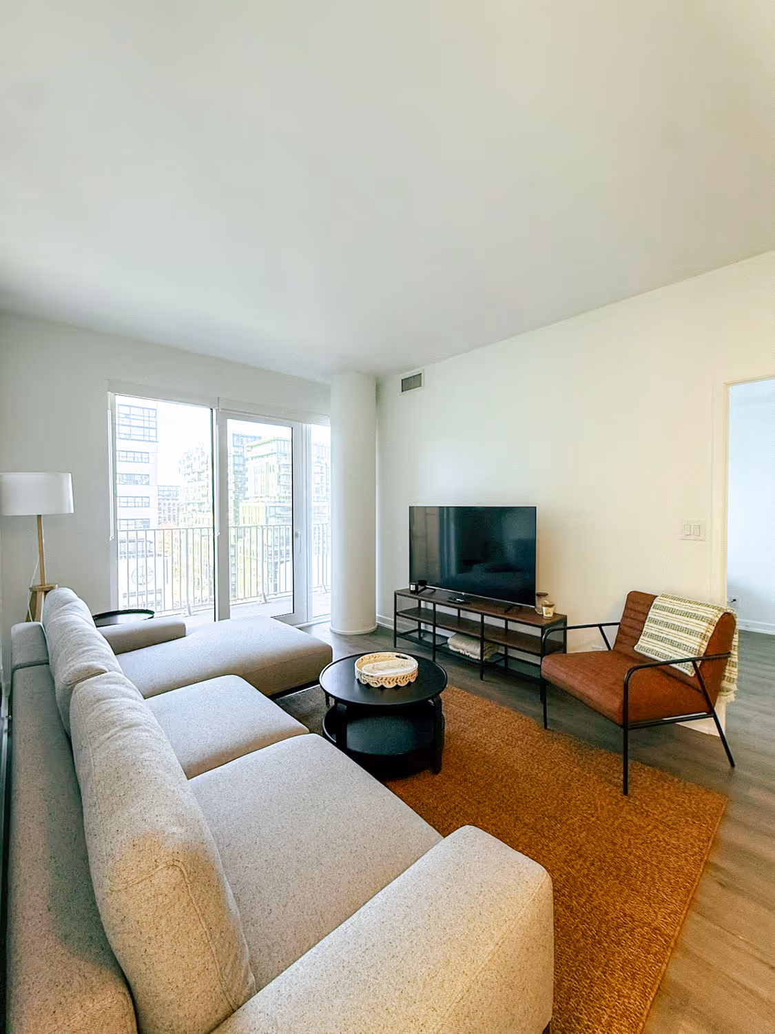 Modern living room with gray sofa, orange armchair, black TV stand, round coffee table, and balcony doors.