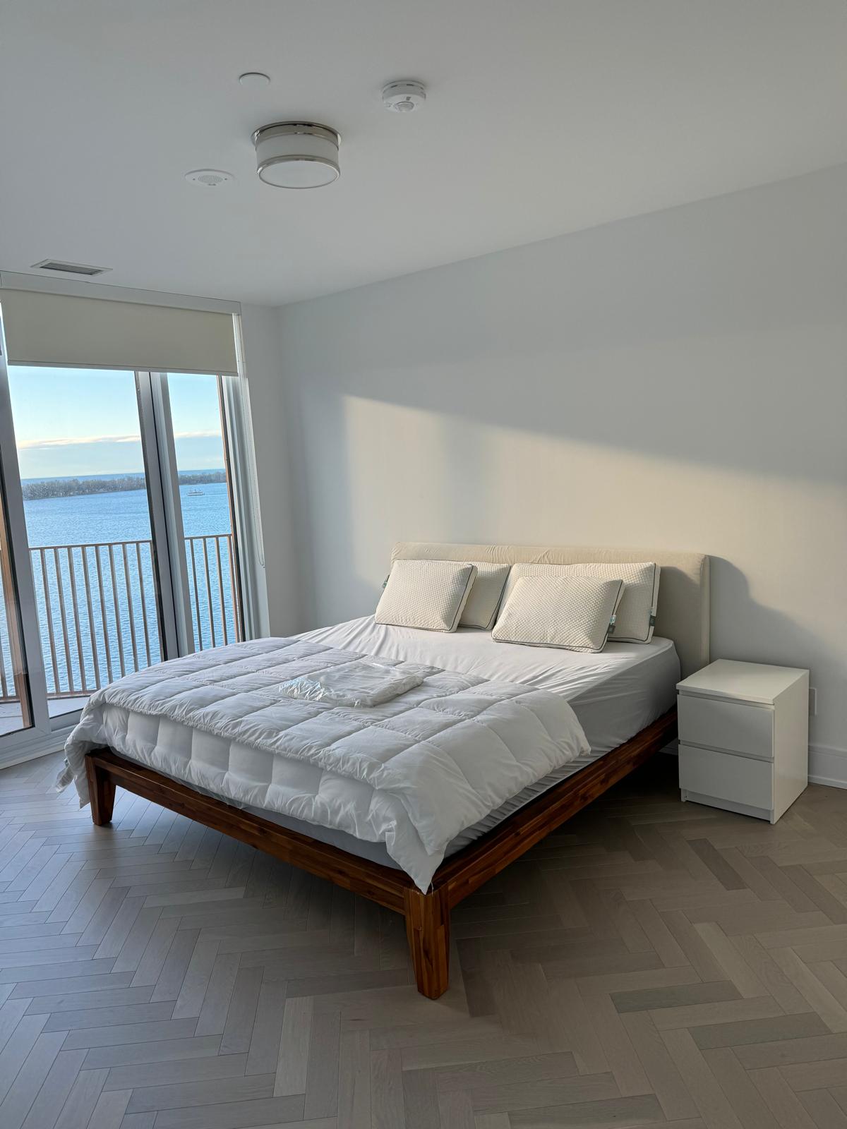 Minimalist bedroom with wooden bed, white bedding, pillows, nightstand, and a water view window.