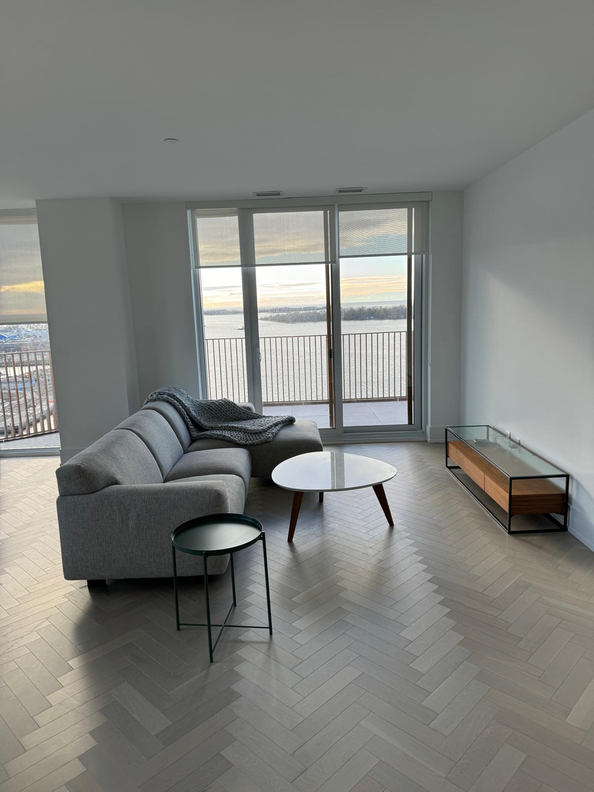 Modern living room with grey sectional sofa, round tables, and large windows overlooking water.