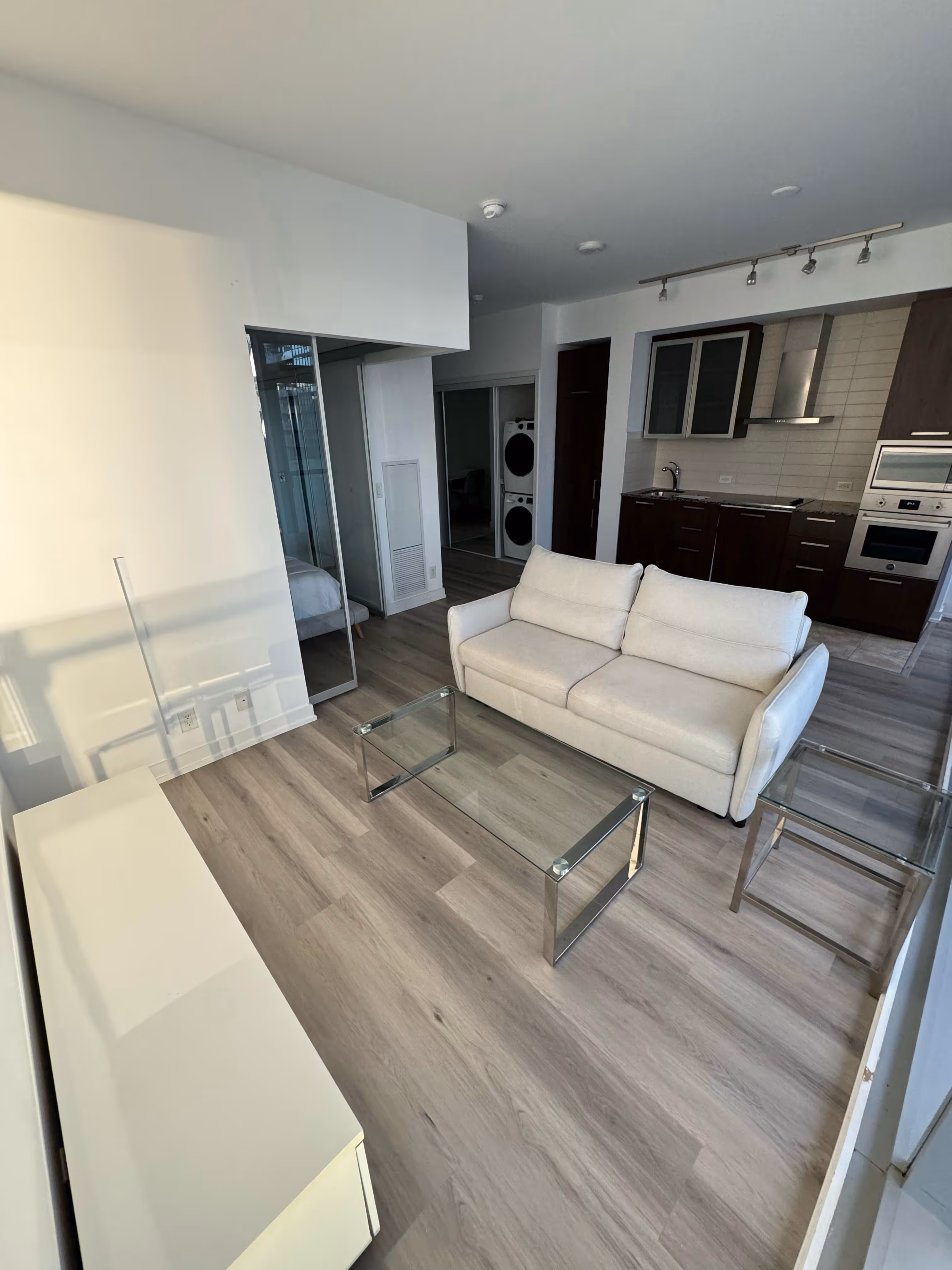 Modern living room with beige sofa, glass coffee and side tables, and kitchen with dark cabinets.