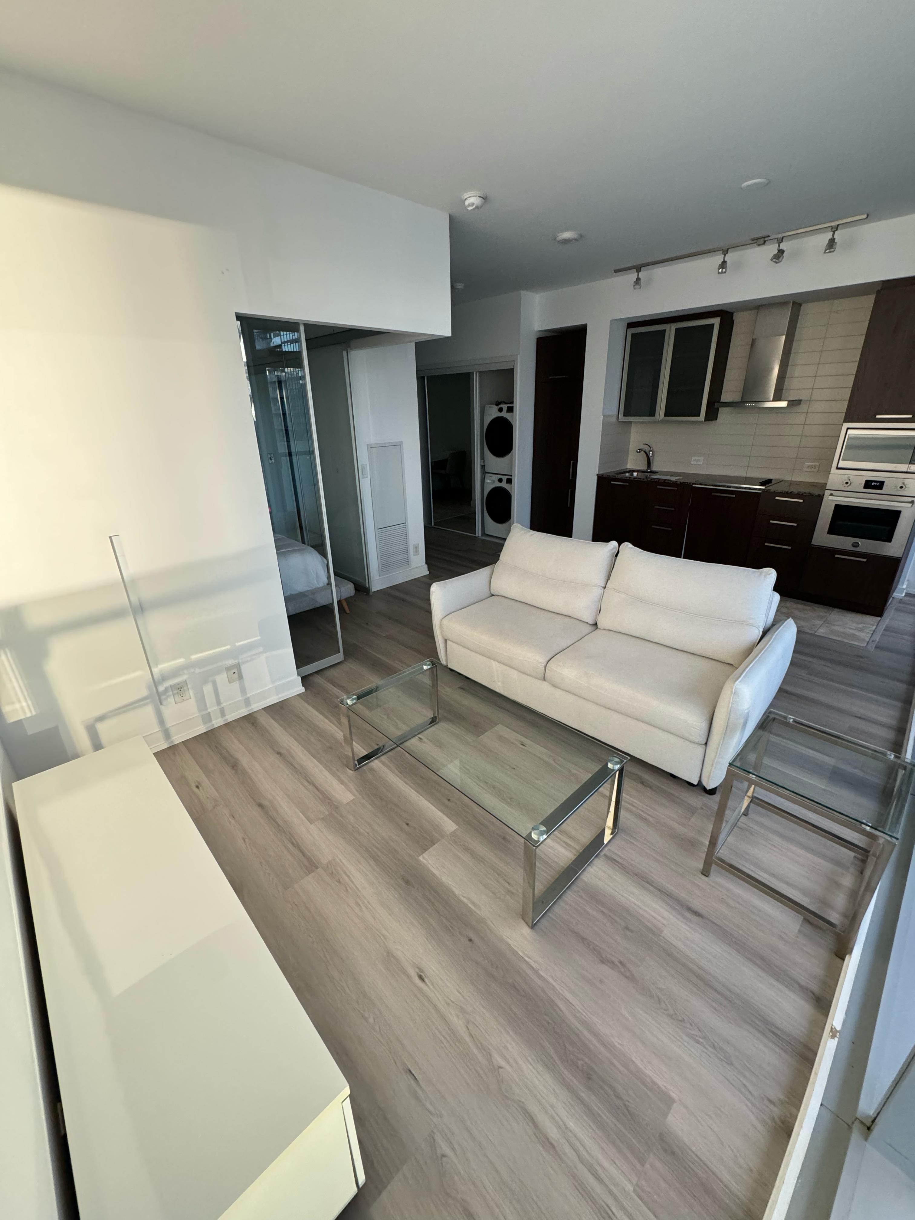 Modern living room with beige sofa, glass coffee and side tables, and kitchen with dark cabinets.