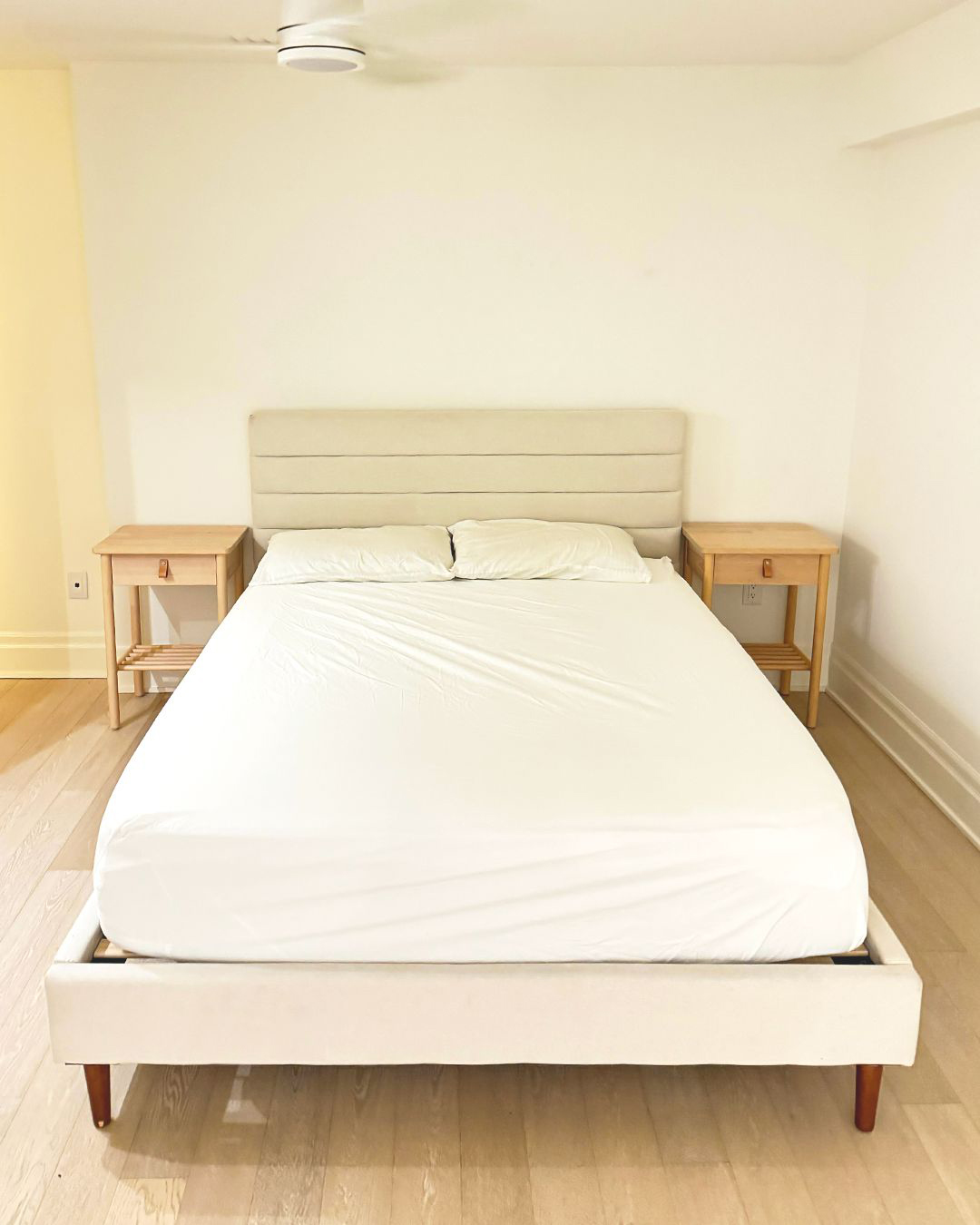 Minimalist bedroom with a beige upholstered bed, white bedding, two wooden nightstands, and light hardwood floors.