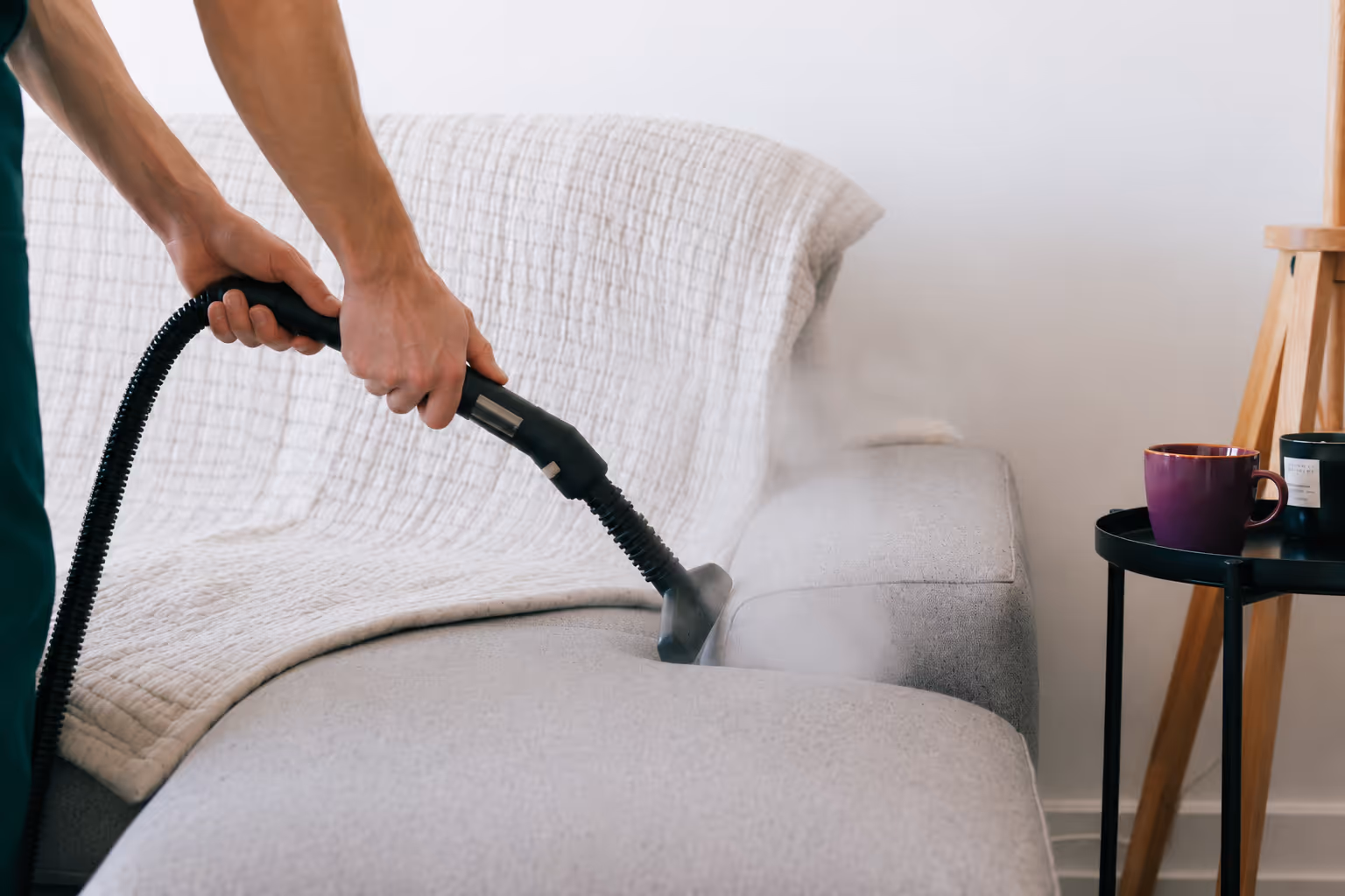 Person steam cleaning a light gray couch with a black hose attachment in a modern living room.